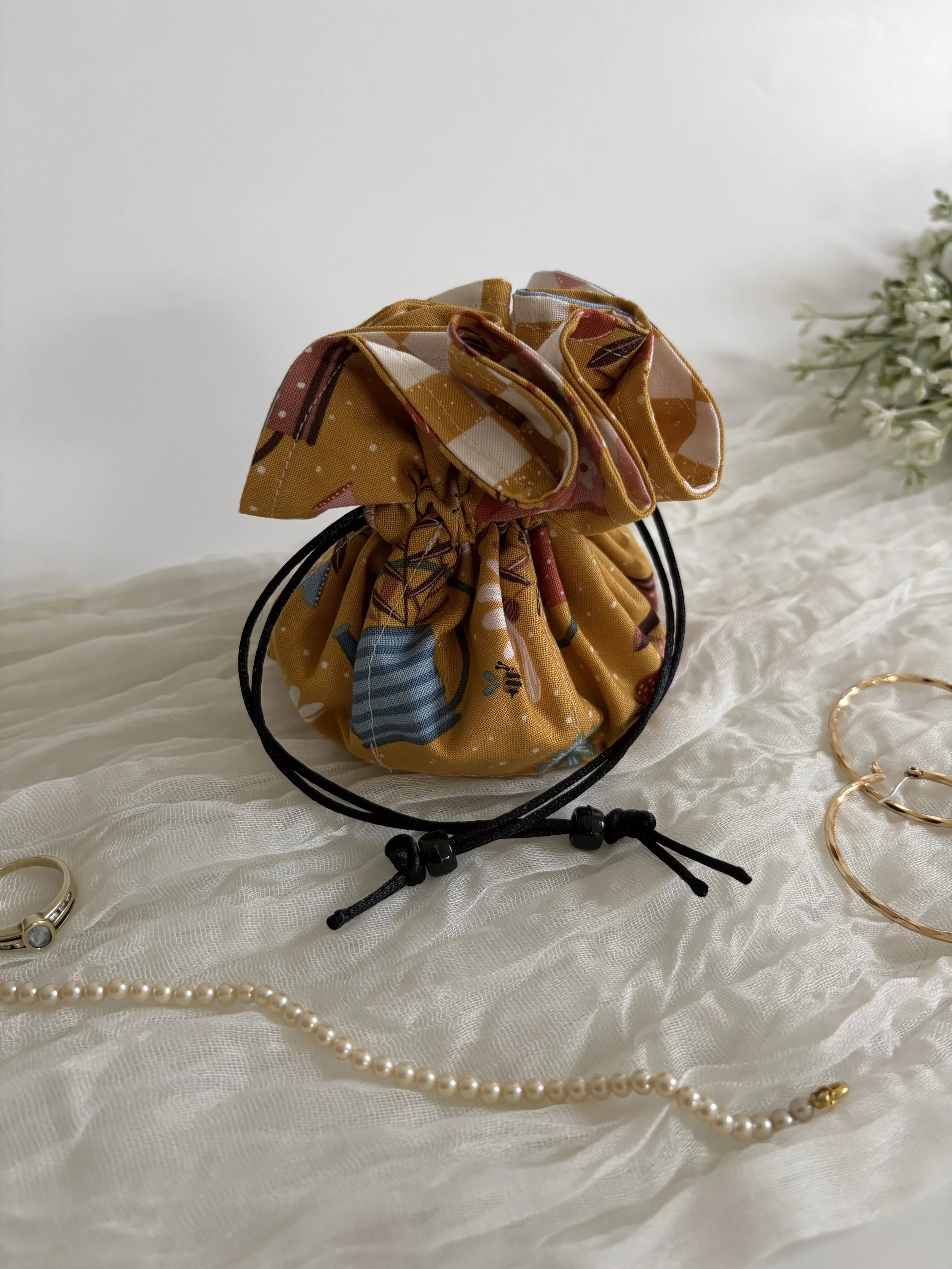 A small, drawstring bag made of fabric with a yellow, red, and blue pattern, on a white textured surface, surrounded by jewelry including gold rings, pearl necklaces, and gold bangles.