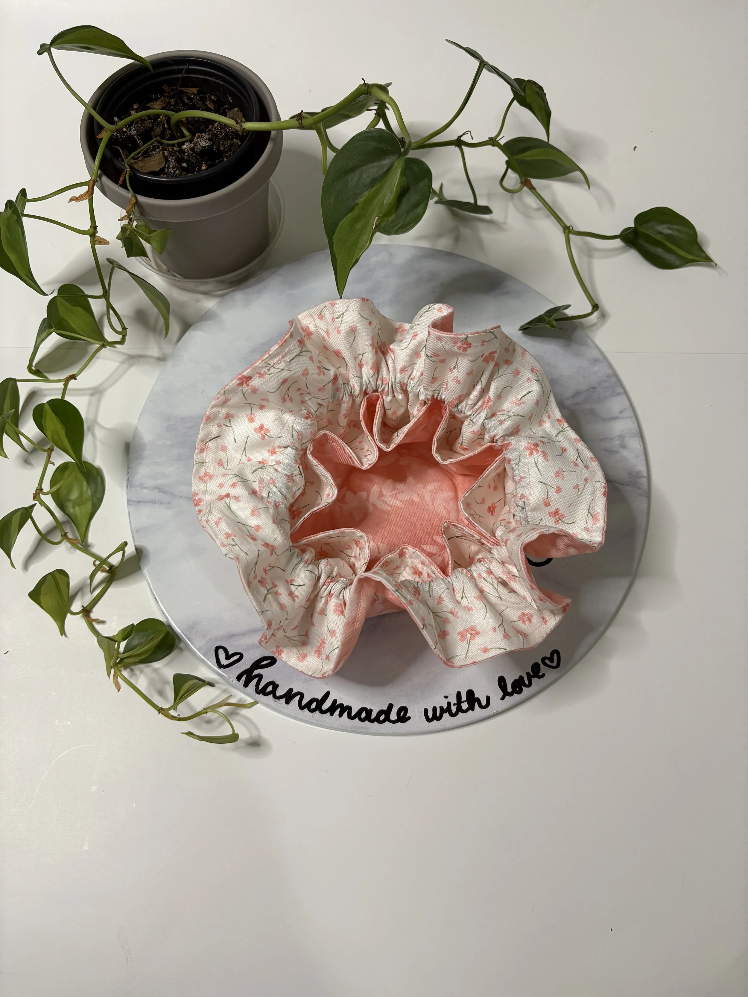 A potted plant with green leaves reclining over a white surface, next to a fabric scrunchie with pink and white patterns. The scrunchie is placed on a round marble tray with a handwritten message 'handmade with love' in black ink and small black hear