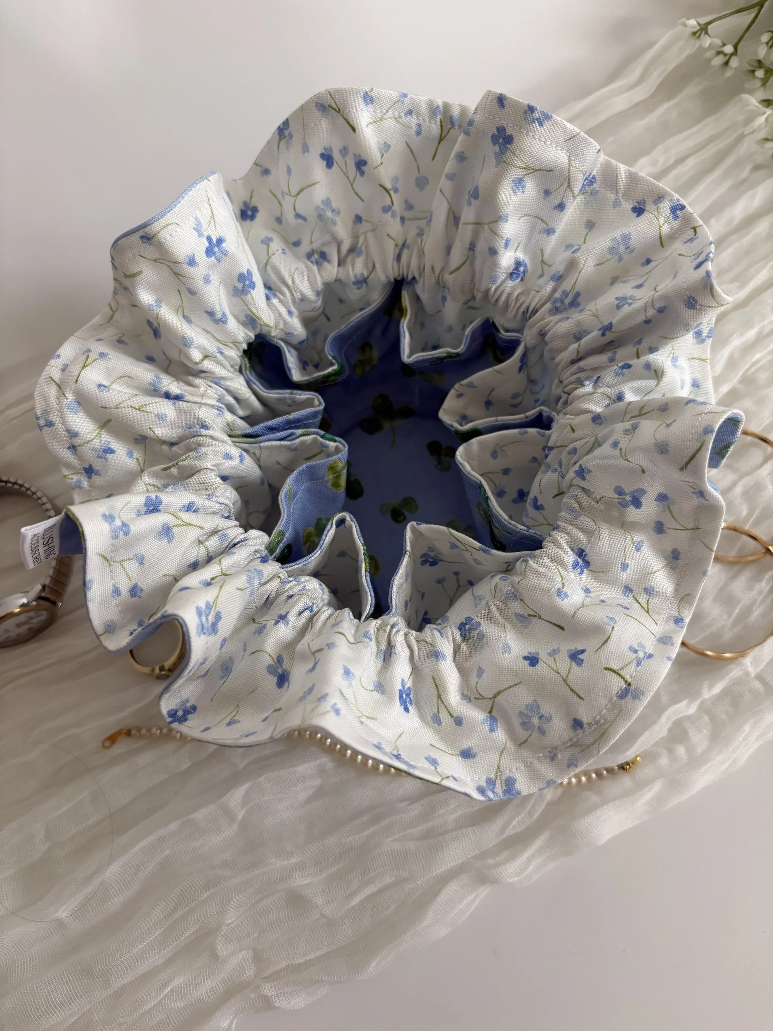 A close-up view of a floral-patterned fabric hair scrunchie with blue flowers on a white background, resting on a white textured surface.