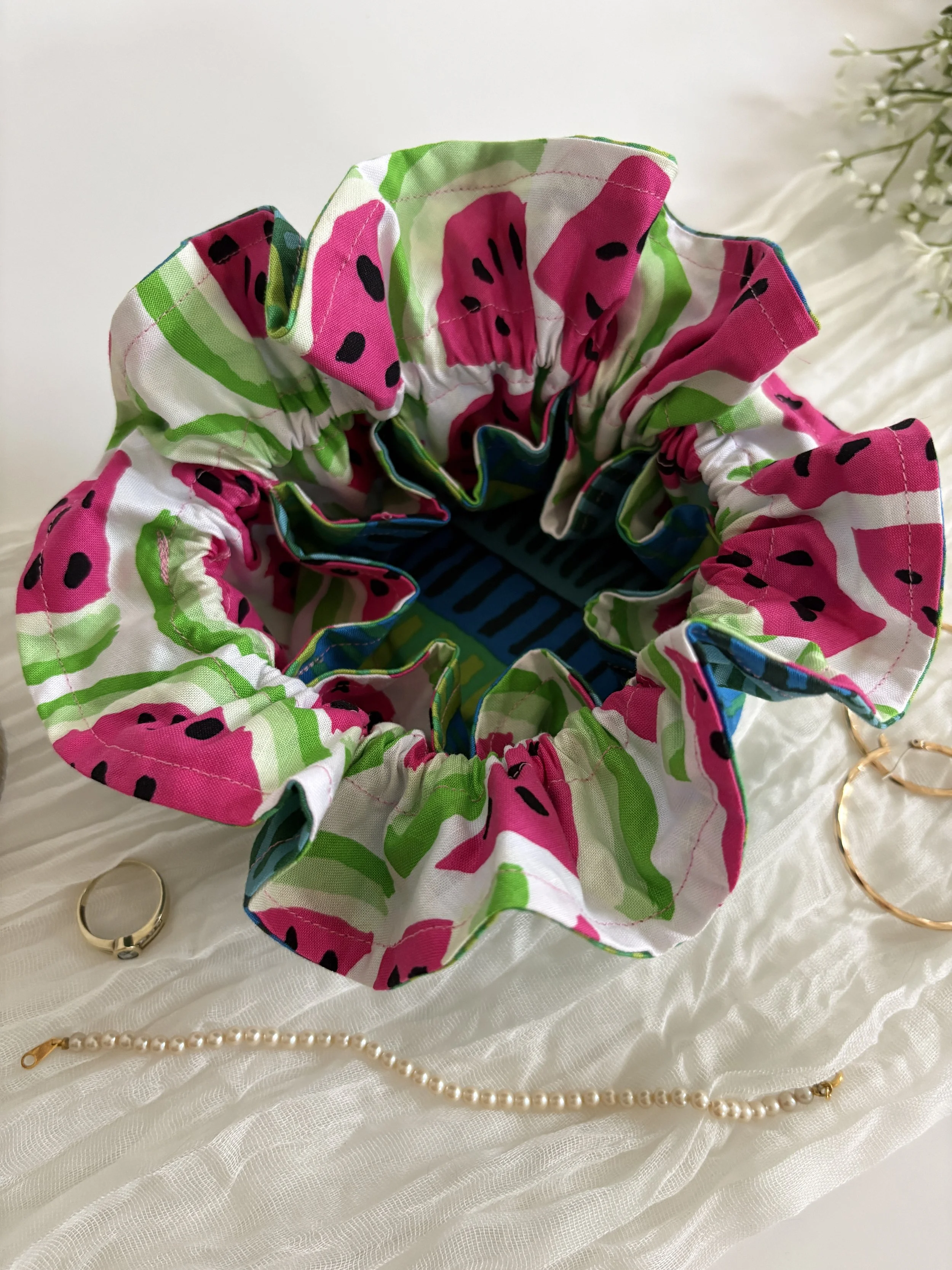 Colorful fabric hair scrunchie with watermelon pattern on white background, gold rings, and pearl necklace.
