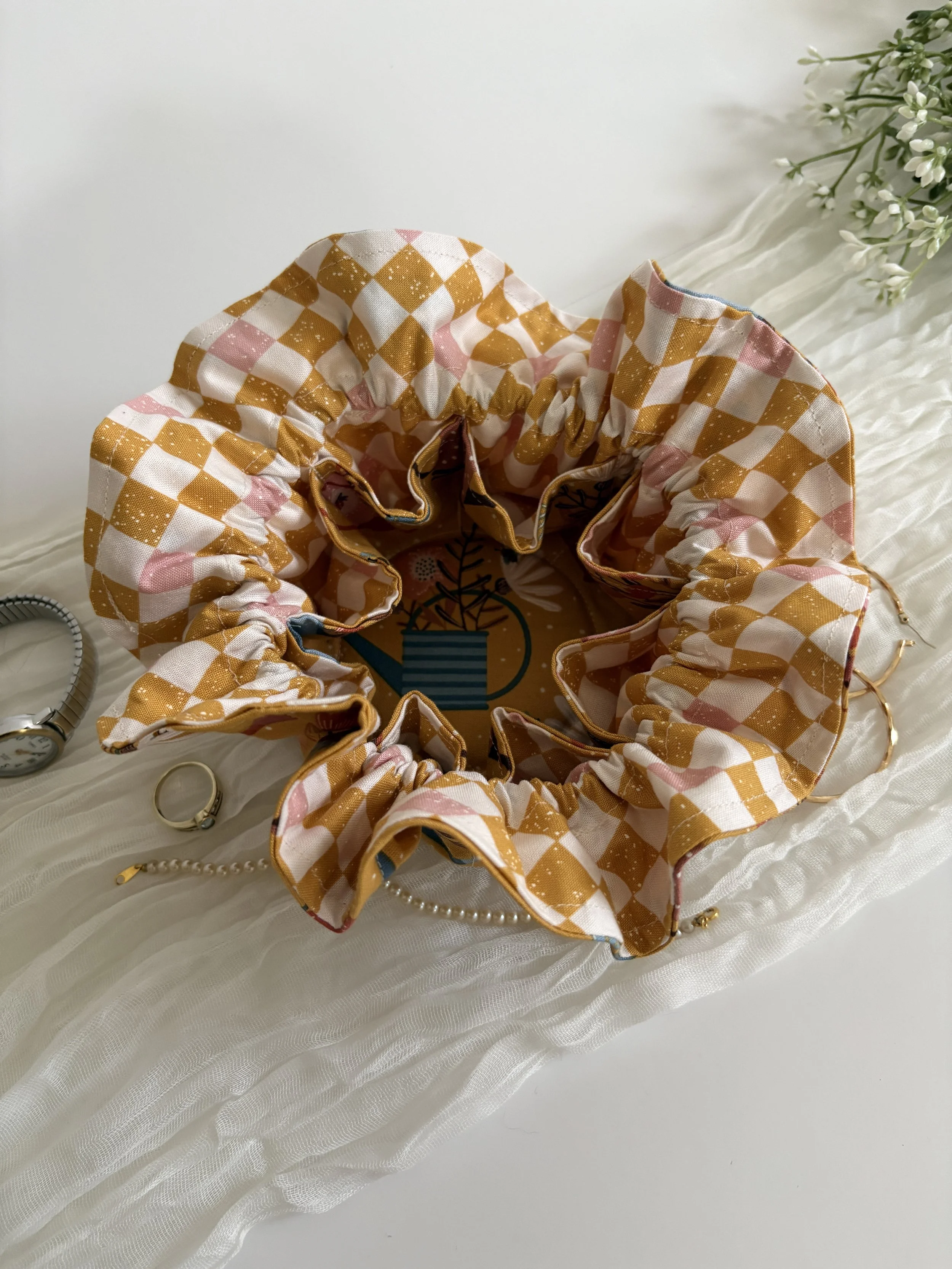 A fabric flower-shaped hair scrunchie with a checkered pattern in beige, pink, and white, placed on a white textured surface with a branch of white flowers nearby.