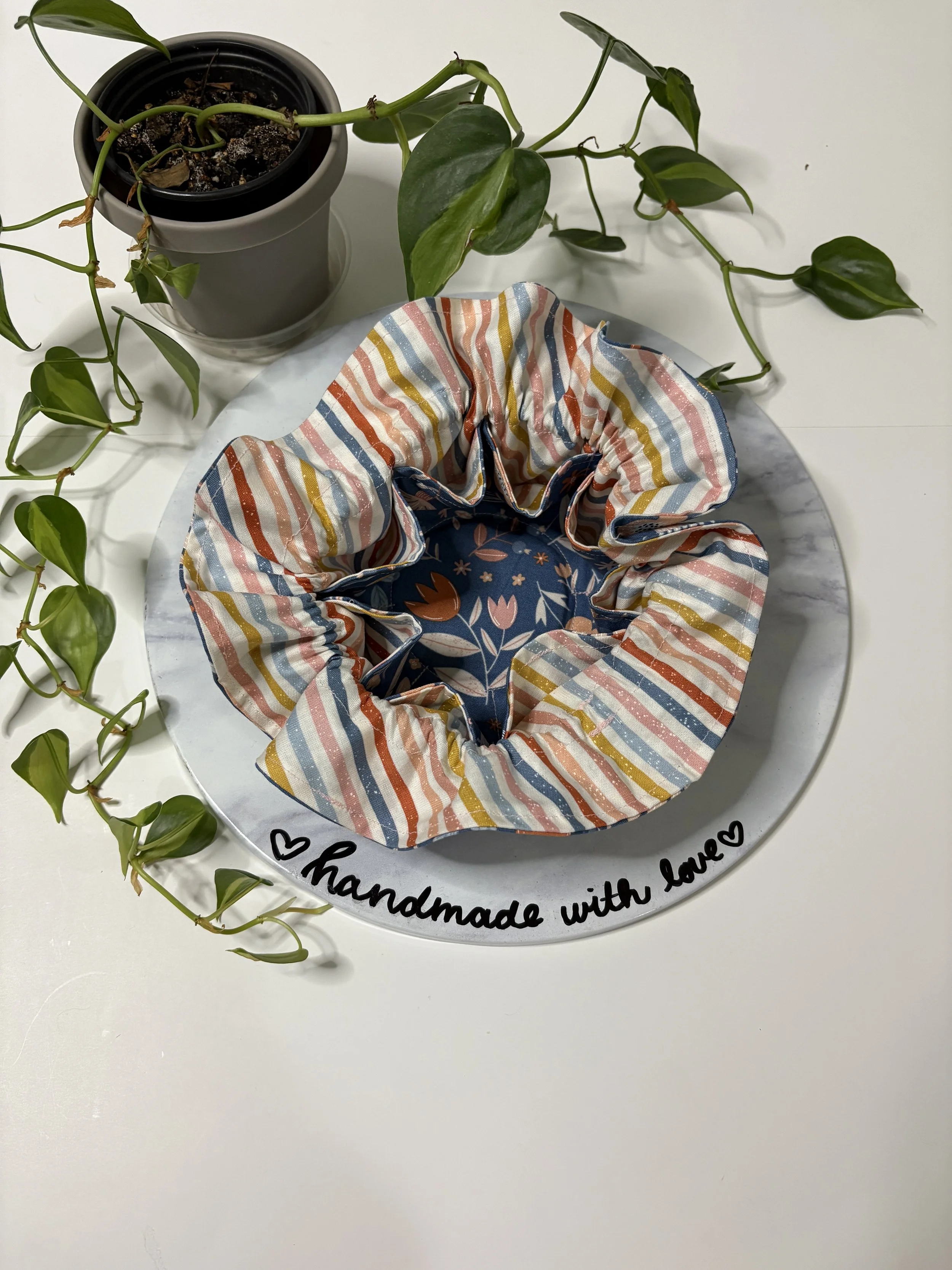 A potted green plant with trailing vines sits on a white table. In front of the plant is a round fabric scrunchie with colorful striped and floral patterns. The scrunchie is placed on a white surface, which has black handwriting reading 'handmade wit