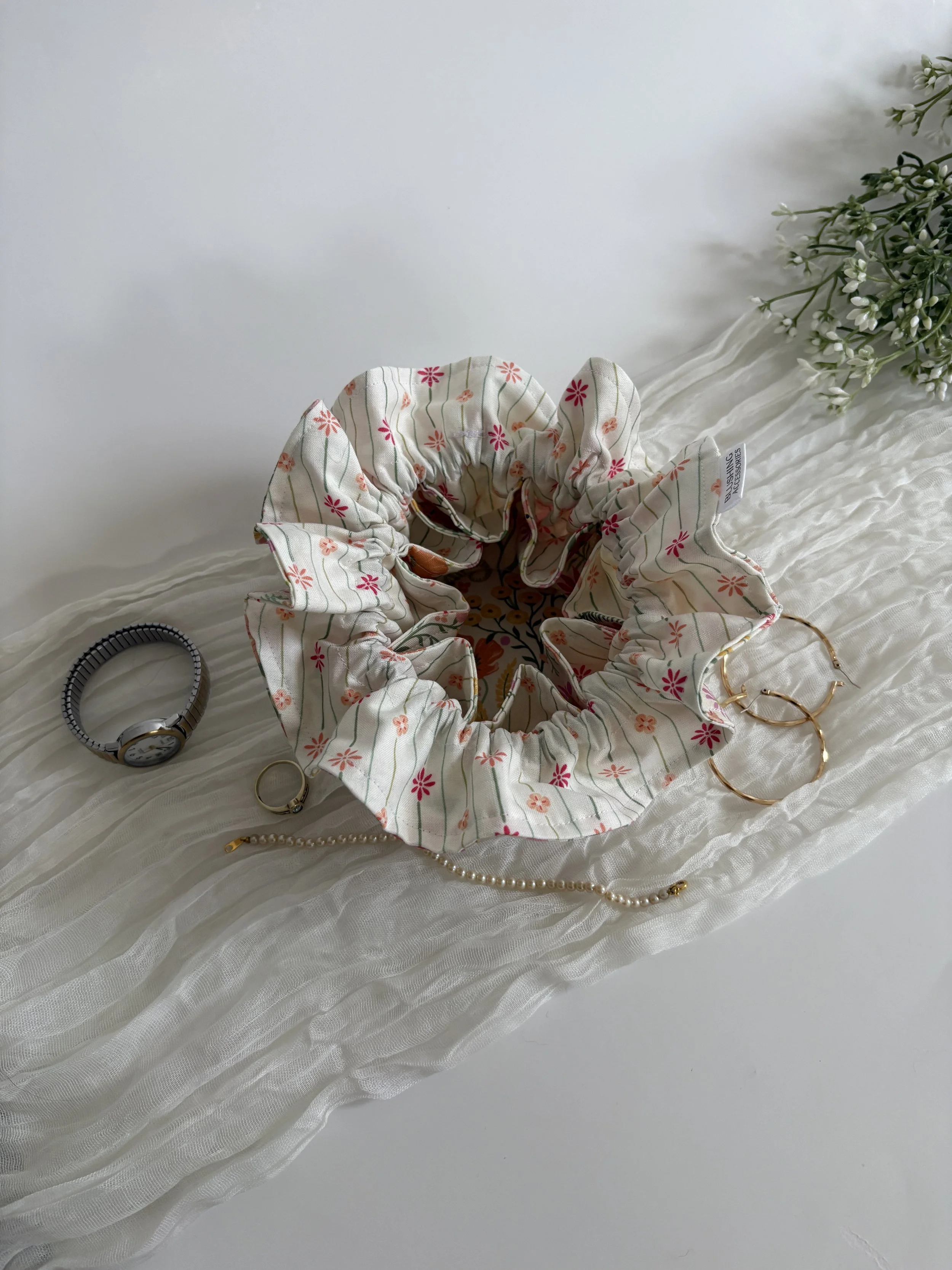 A floral scrunchie, a pearl bracelet, a gold bracelet, a black hair tie, and a pair of gold hoop earrings are arranged on a white textured fabric surface with a small bouquet of white flowers in the upper right corner.