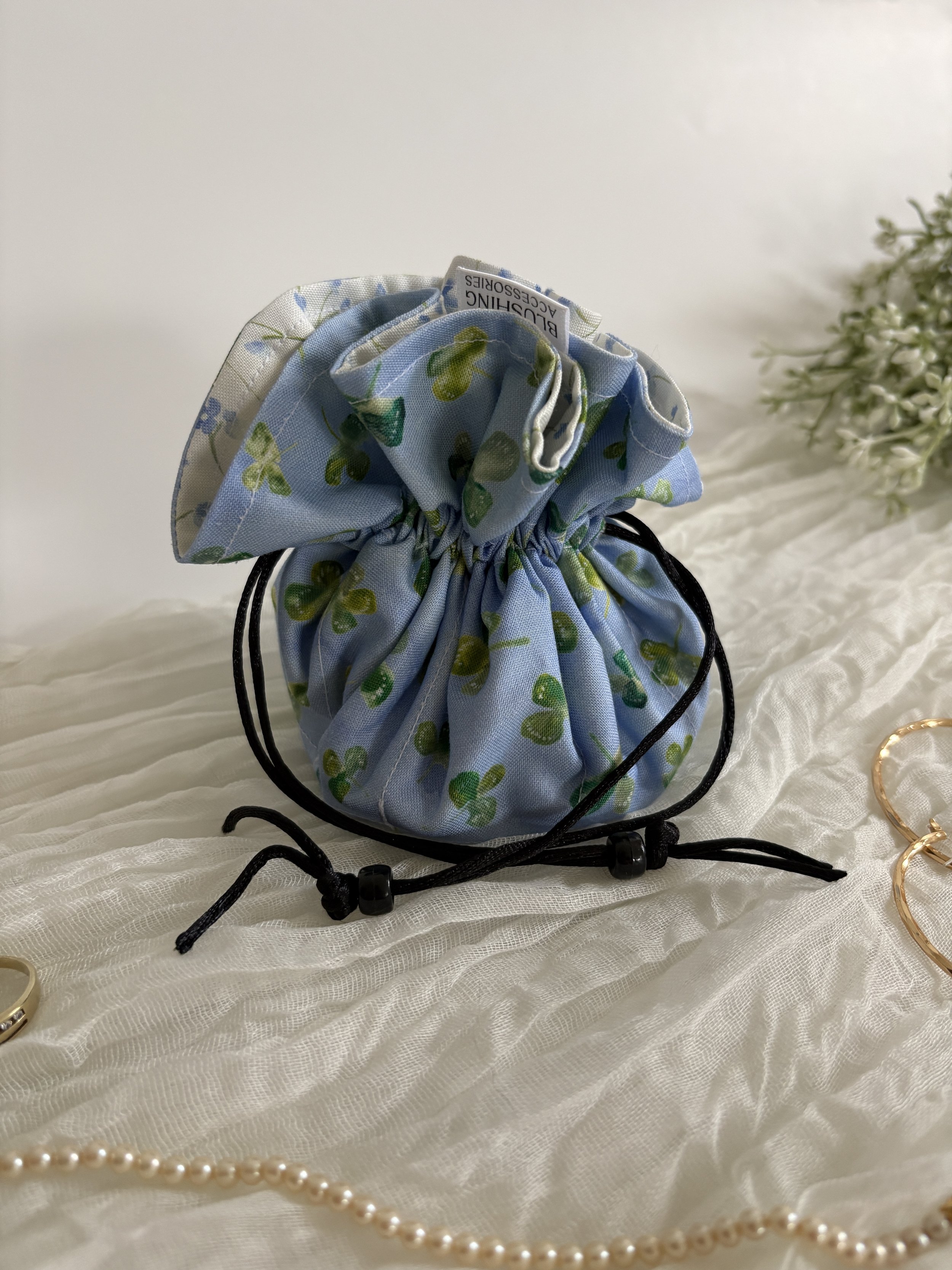 Blue fabric pouch with green clovers, gathered at the top with a drawstring, on a white satin surface, with pearl necklace and gold bracelet nearby.