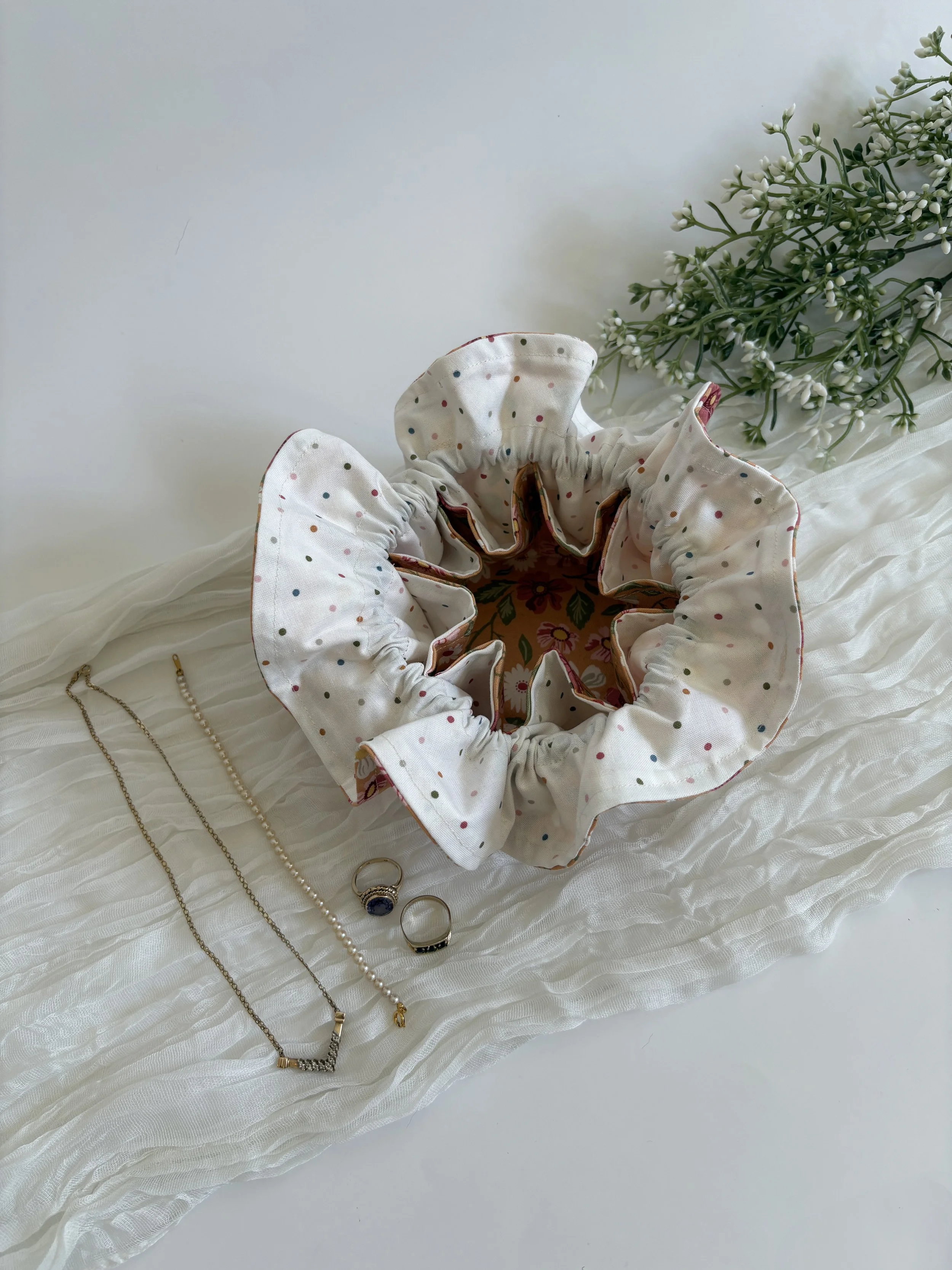 A flat lay of jewelry items including a pearl necklace, a ring, and a bracelet placed on white fabric with a sleeveless floral top, cream-colored with multicolored polka dots, laid upside down. A bouquet of white flowers is on the right side of the floral top.