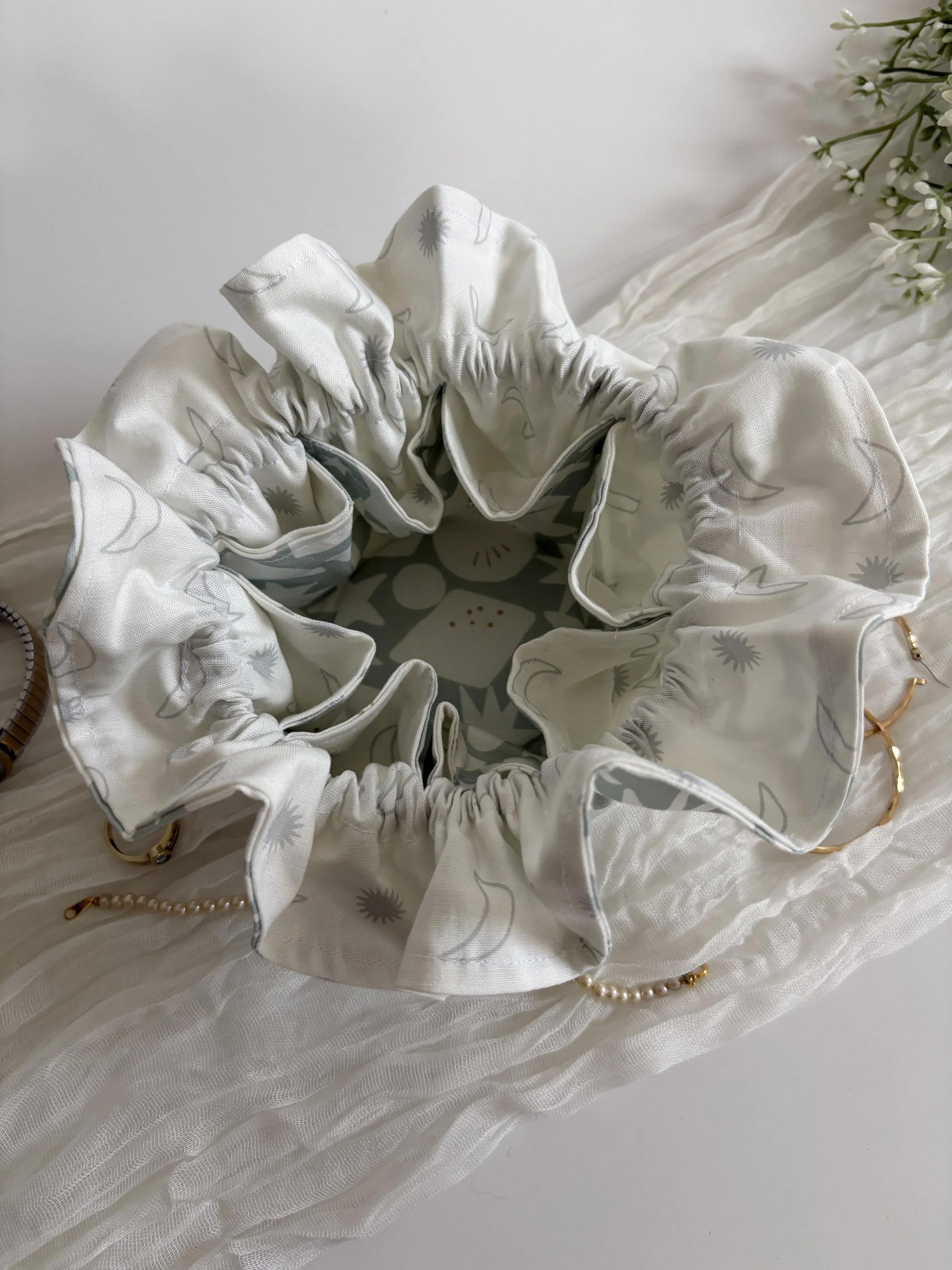 Close-up of a white fabric headband with a ruched design and a delicate flower pattern, resting on a textured white surface, with a small segment of a pearl necklace visible.