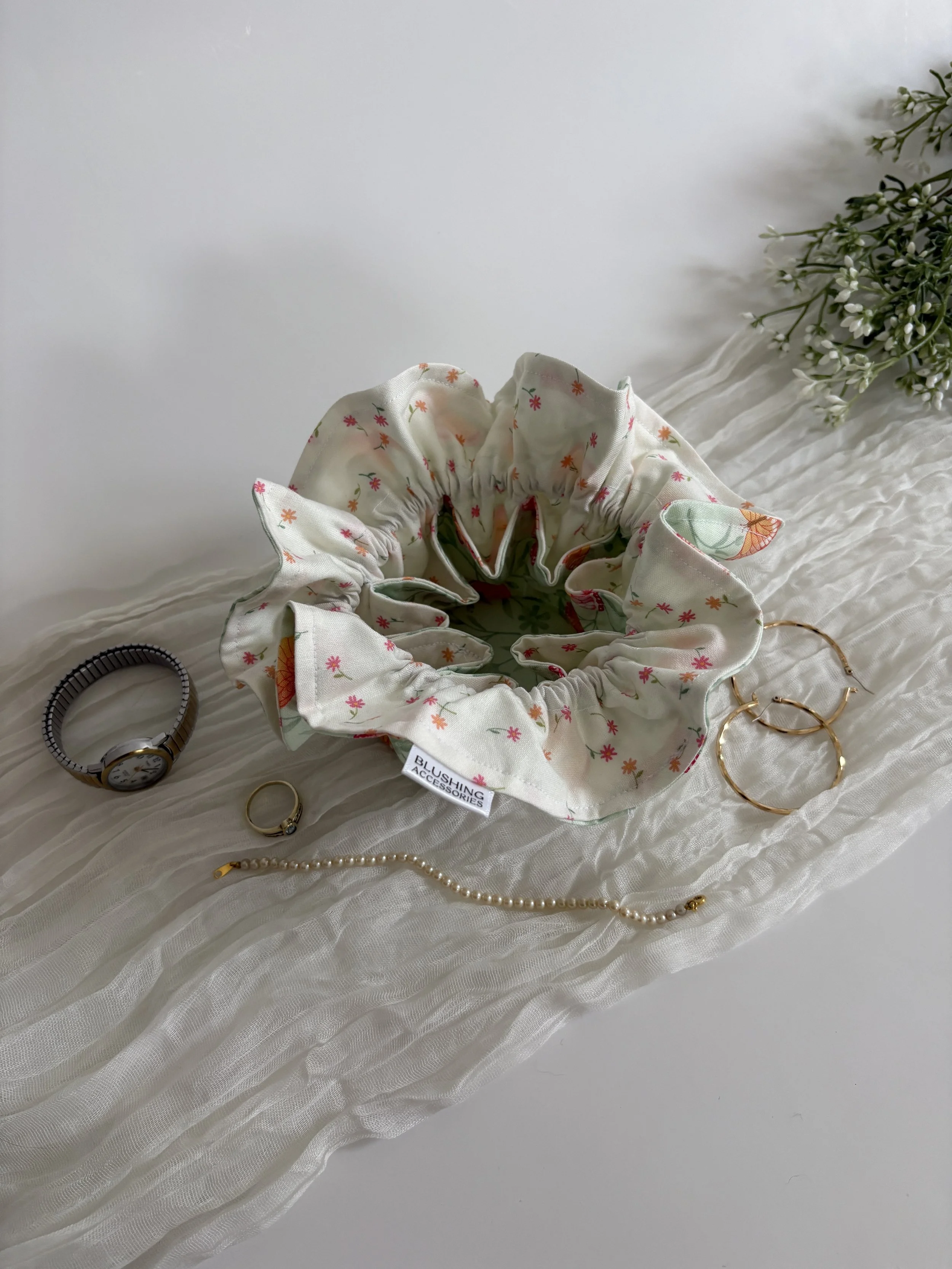 A collection of jewelry including a black and gold watch, a pearl bracelet, and gold hoop earrings, arranged on a white textured fabric next to a fabric hair scrunchie with a floral pattern, and a bunch of white flowers with green leaves.