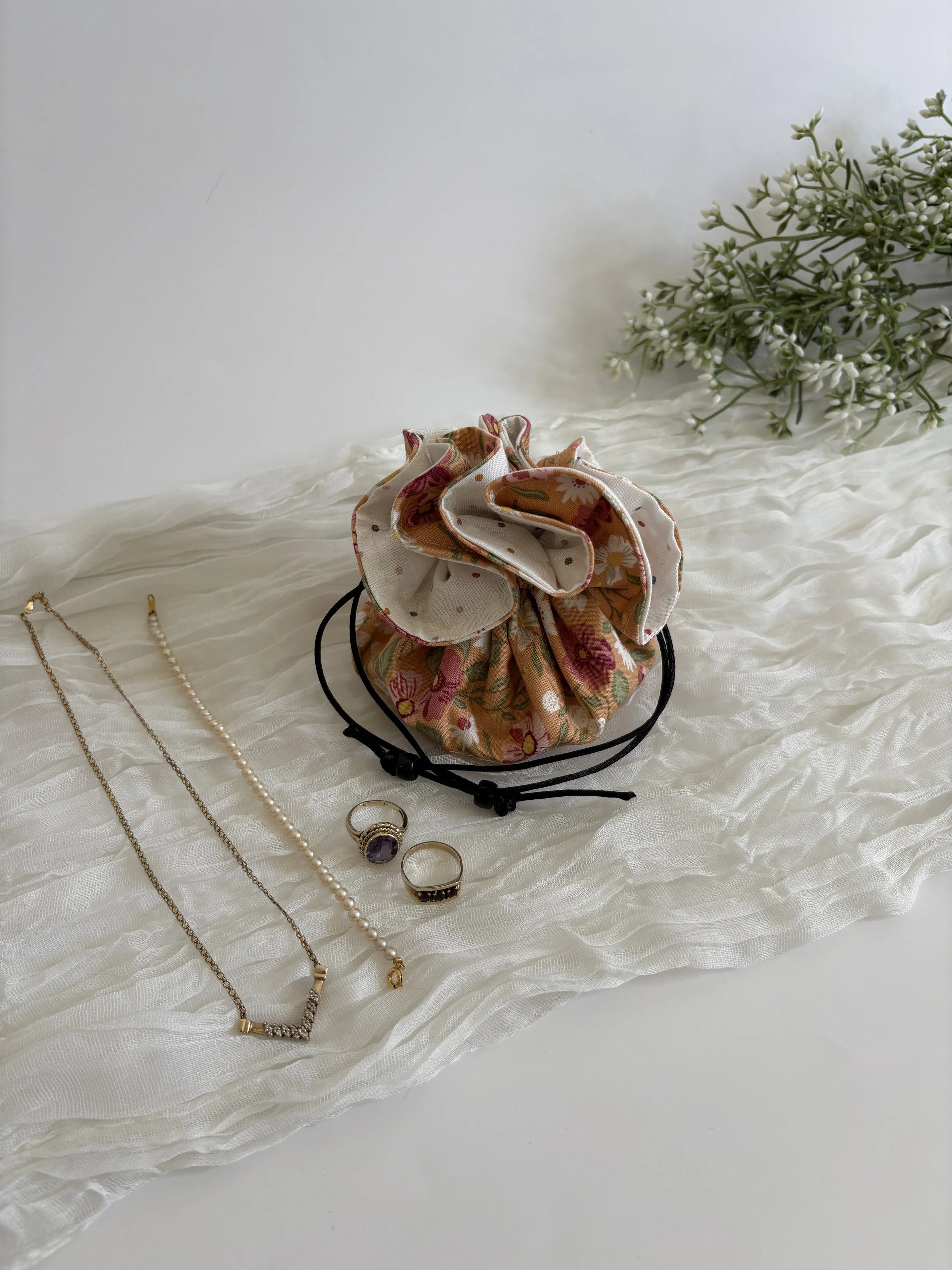 Jewelry including necklaces, rings, a scrunchie, and a bracelet displayed on textured white fabric with a bouquet of white flowers in the background.
