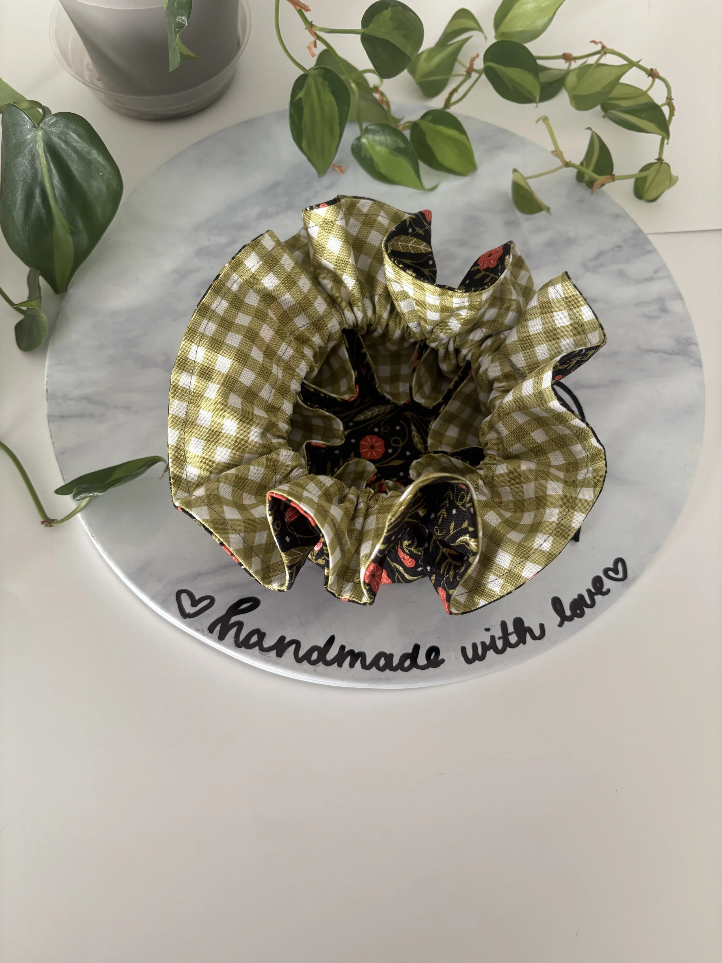 A round marble tray with the words 'handmade with love' written around the edge. On the tray, there is a fabric scrunchie with a checkered pattern in beige and white, and part of a black fabric with red and gold floral designs. In the background, the
