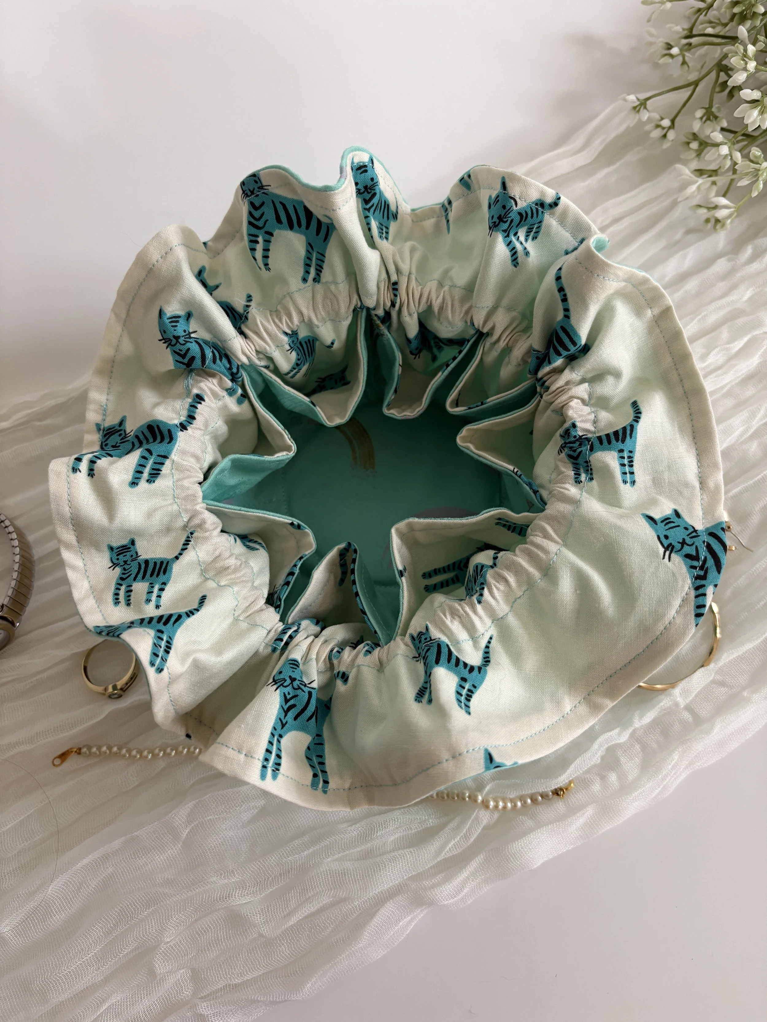 A fabric scrunchie with blue tiger print rests on a white textured surface next to a string of pearls and a small cluster of white flowers.