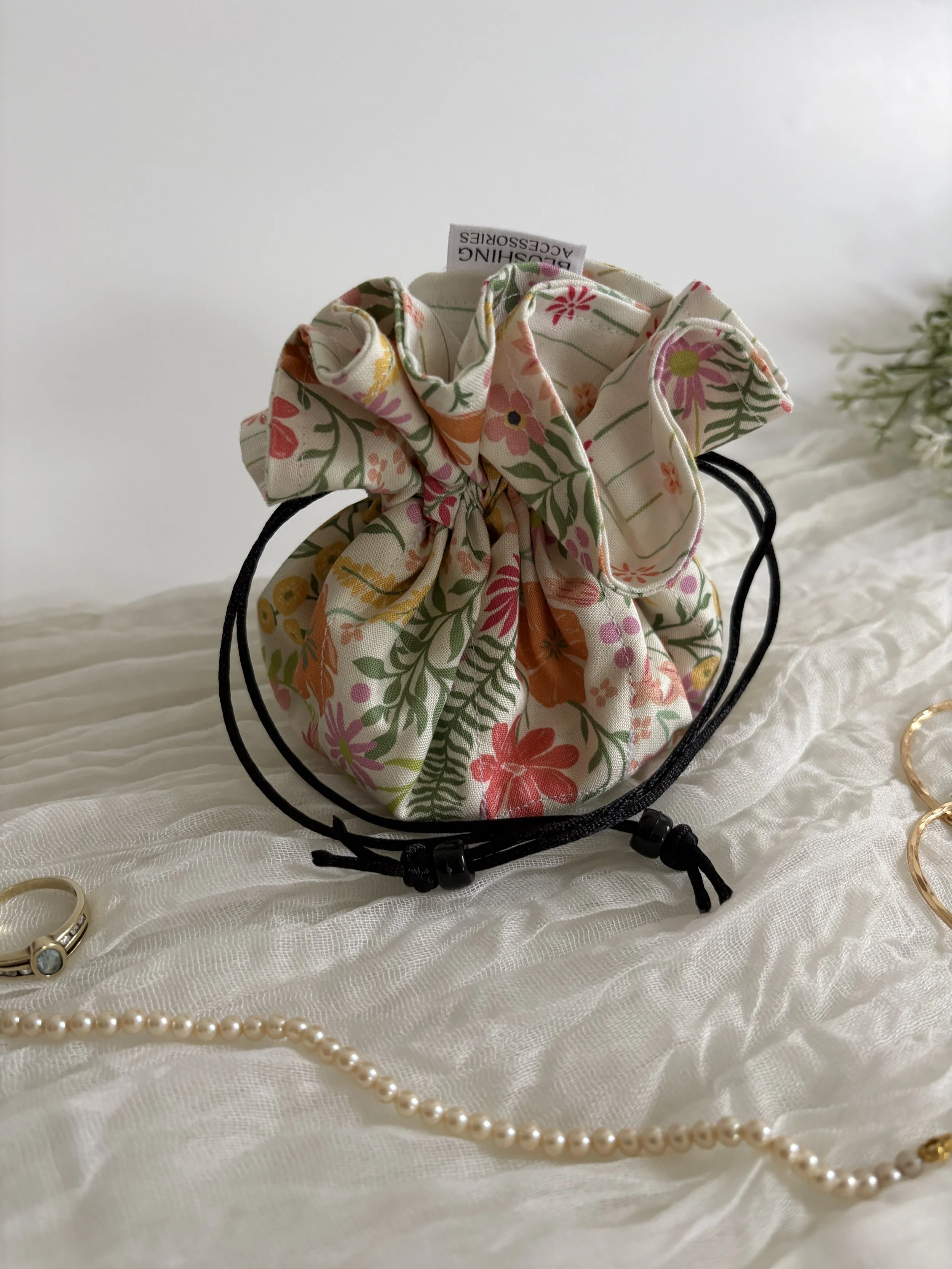 A floral fabric scrunchie with a tag reading "Dressing Accessories" on a white textured fabric, surrounded by jewelry including a pearl necklace, gold rings, and bracelets.