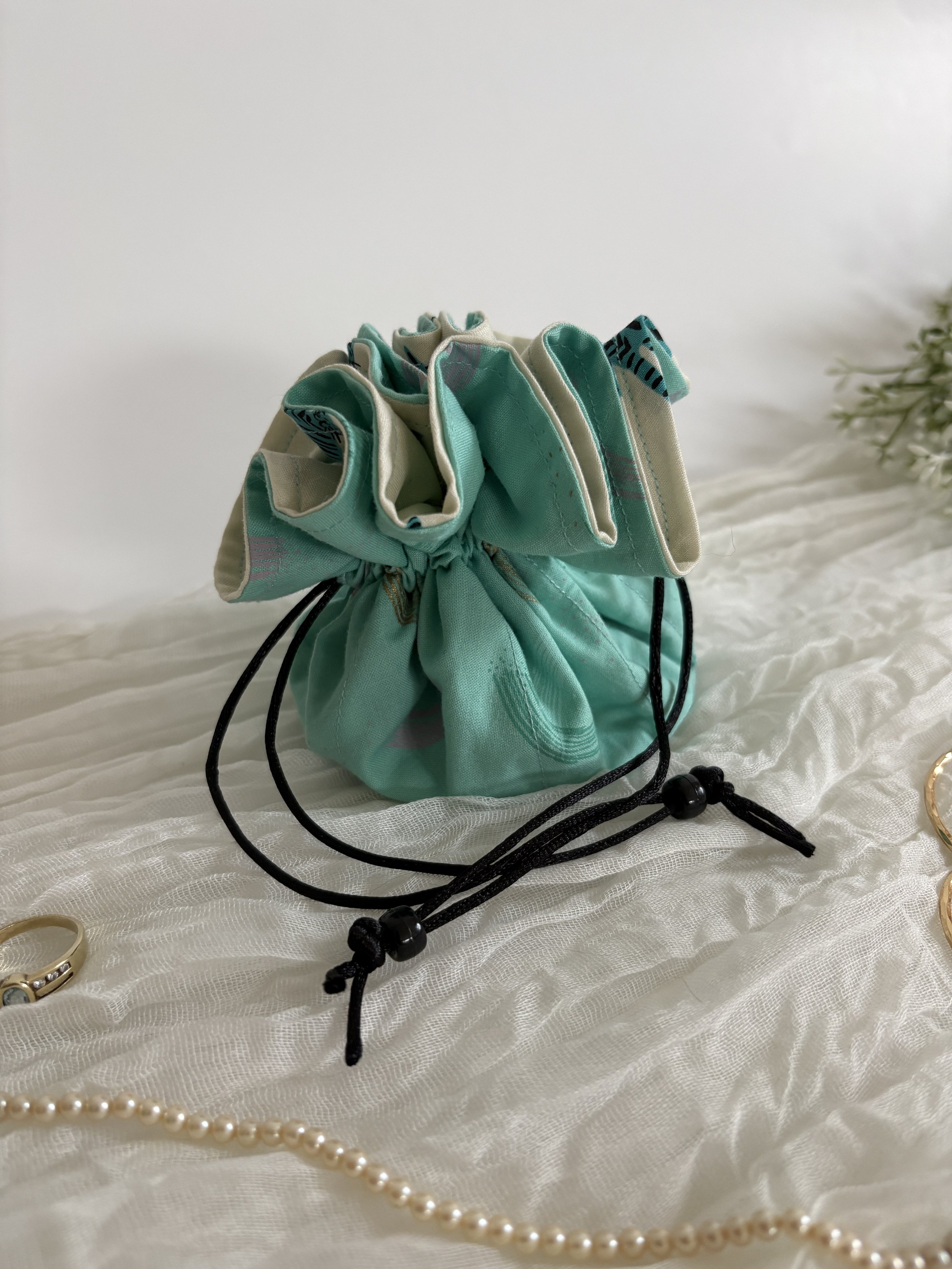 A teal and cream fabric drawstring pouch with a bow, placed on a white textured surface. Partially visible jewelry including a pearl necklace, a ring, and a gold bracelet are also on the surface.