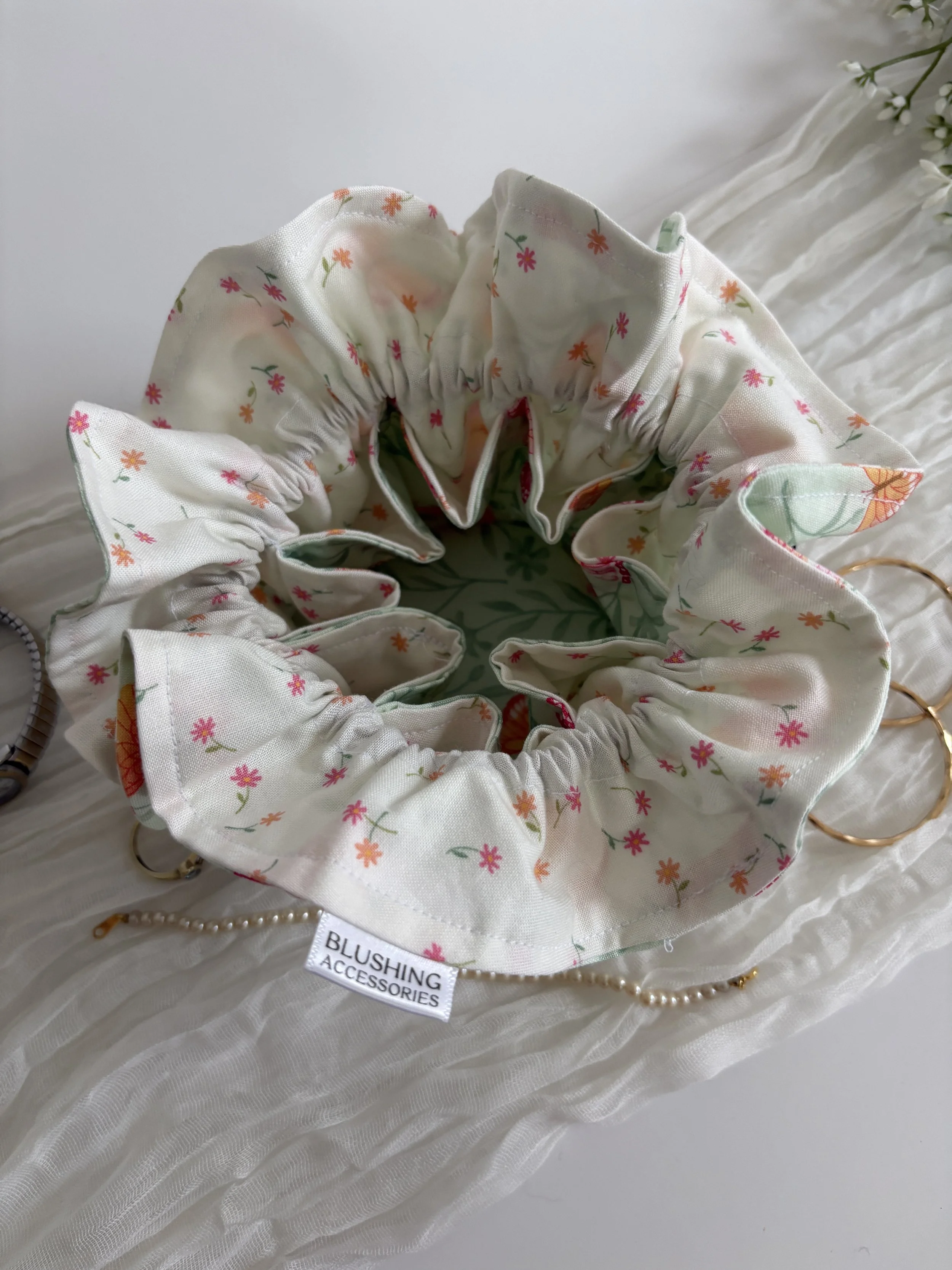 A floral-printed scrunchie, a pearl necklace, and a ring on a white fabric surface with a small label reading 'Blushing Accessories'.