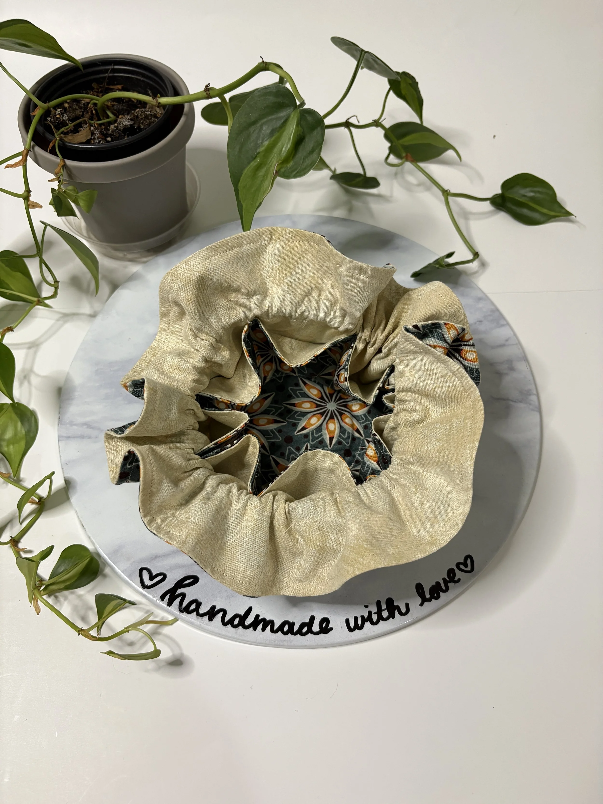 A potted vine with heart-shaped green leaves and a fabric drawstring bag with black, orange, and white pattern inside. The items are on a white table with a round marble board that has a handwritten message saying 'handmade with love' and hearts.