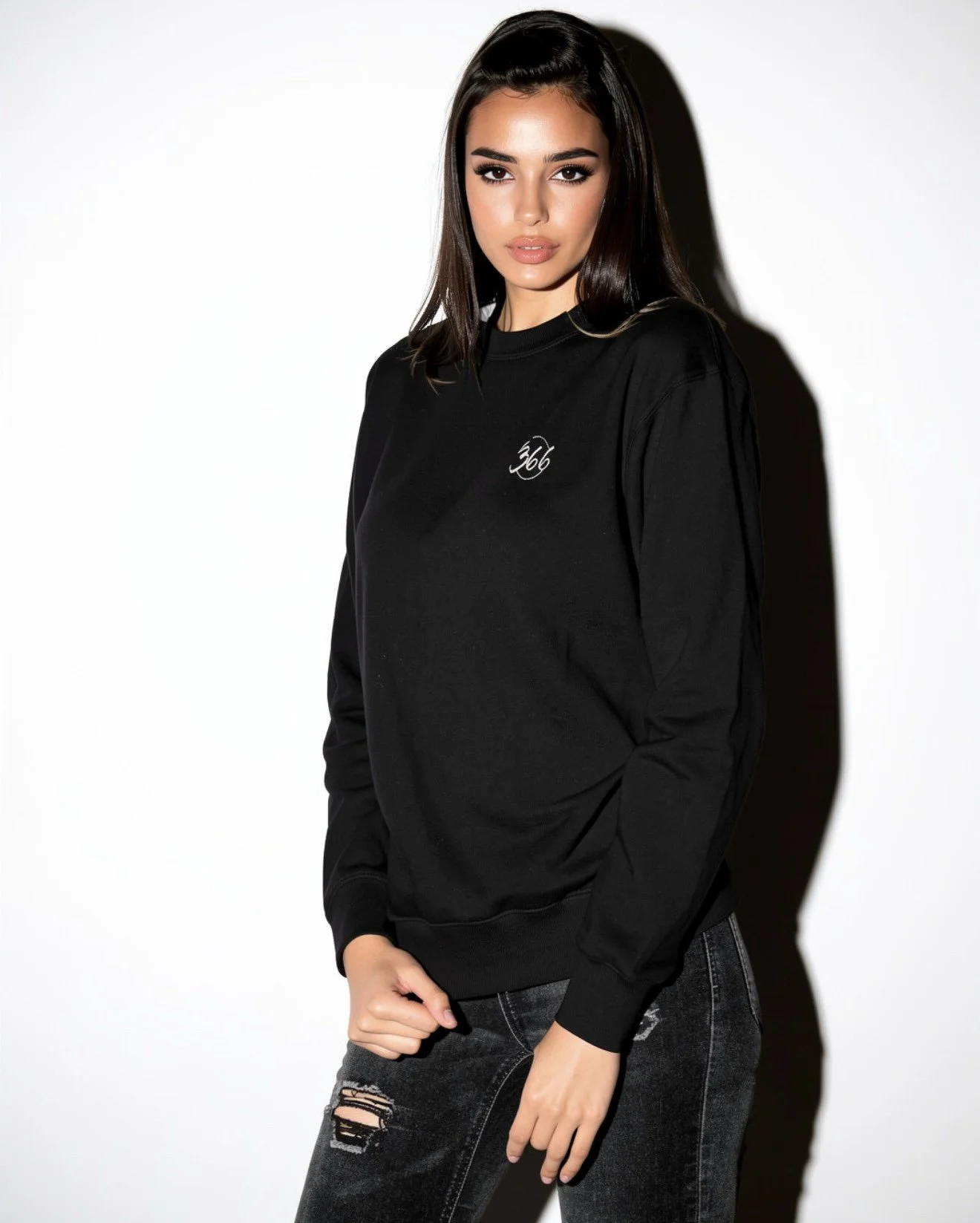 A young woman with shoulder-length brown hair and makeup, wearing a black sweatshirt with a small design on the chest, black ripped jeans, standing with her left arm relaxed and her right hand holding her waist, against a plain white background.