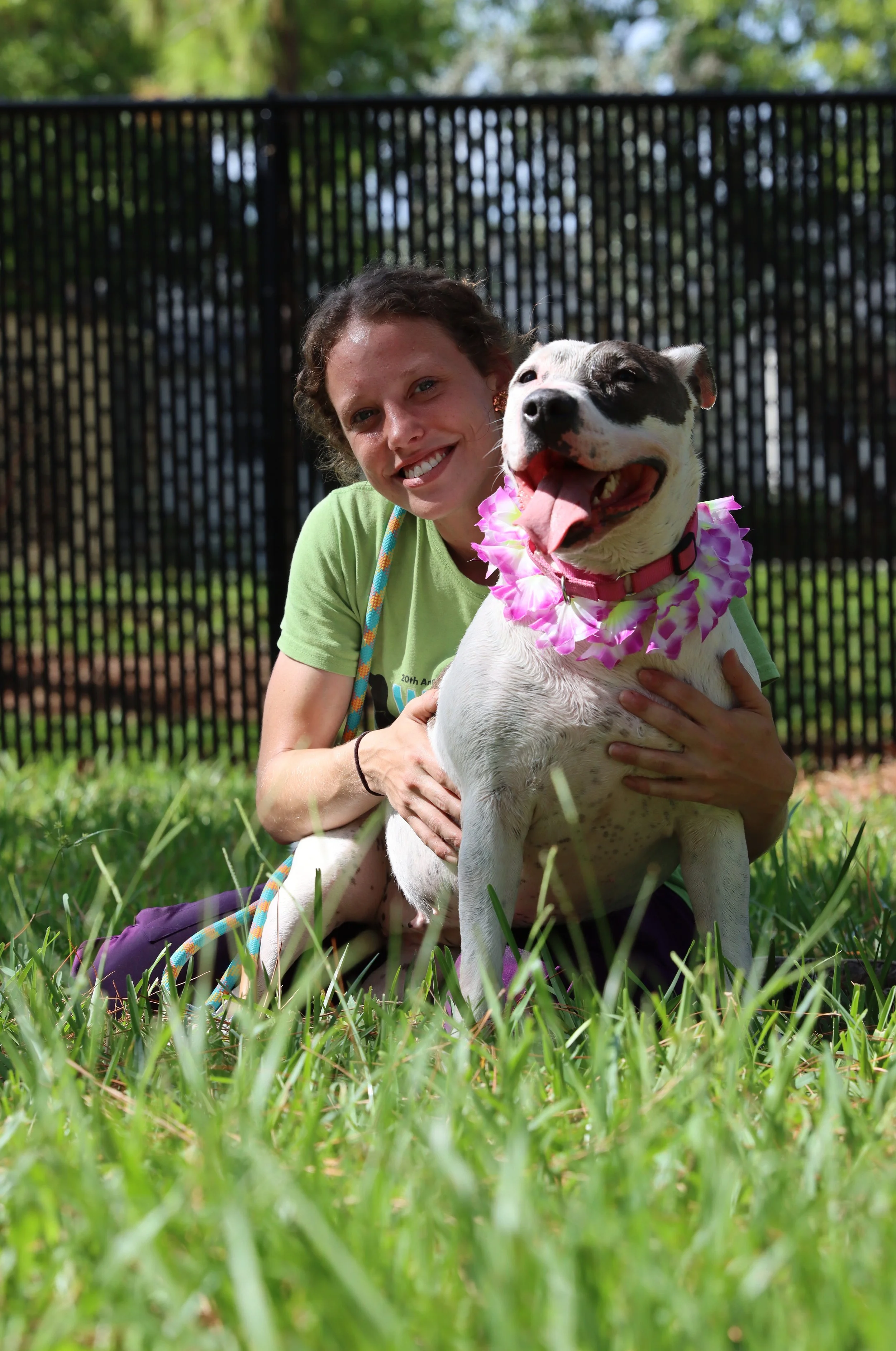 A young woman smiling and hugging a happy dog with a pink lei around its neck in a grassy outdoor area