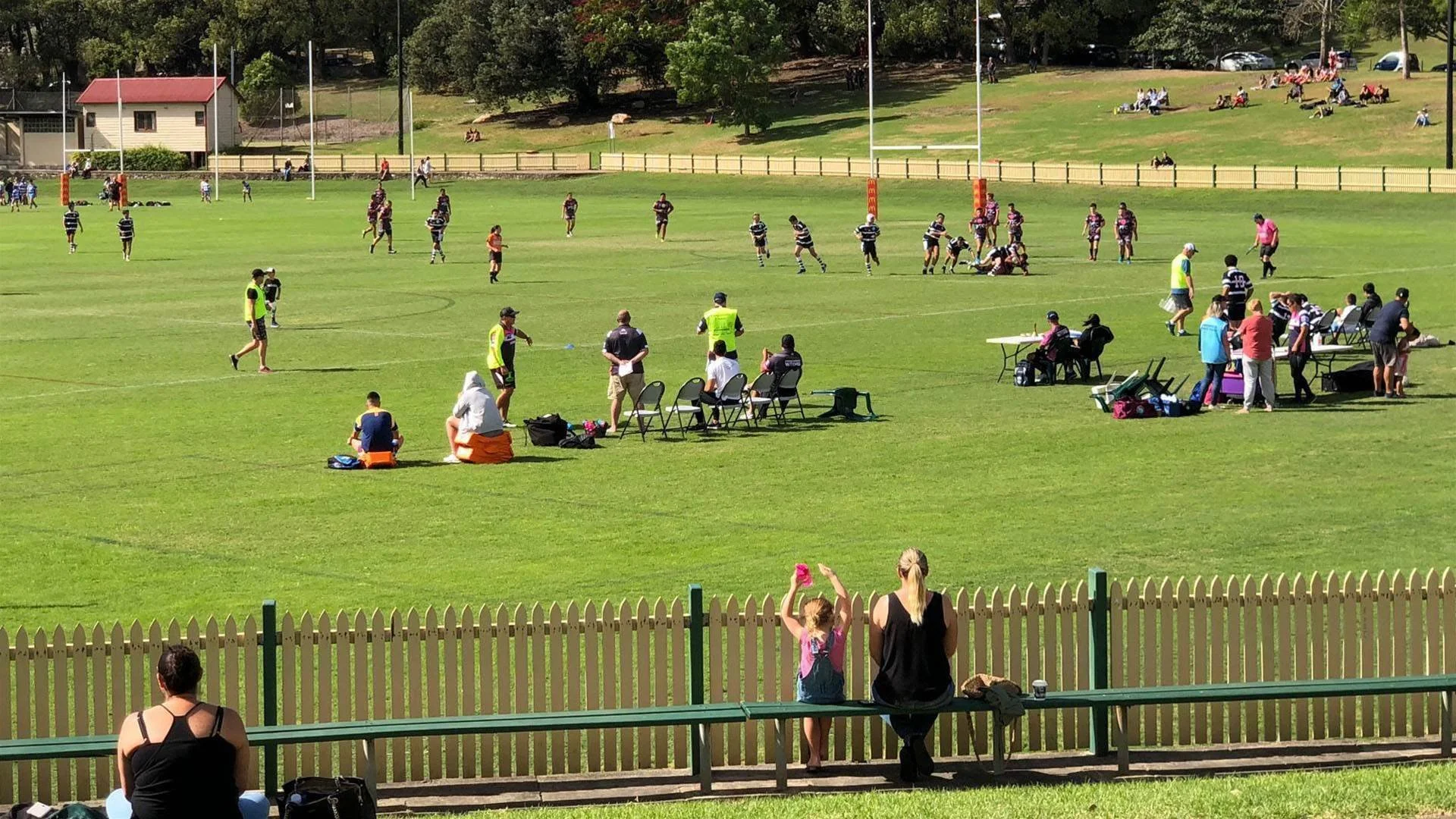 Birchgrove Oval with balmain junior rugby league players, spectators sitting on benches and standing, and people watching from a distance on a grassy hill.