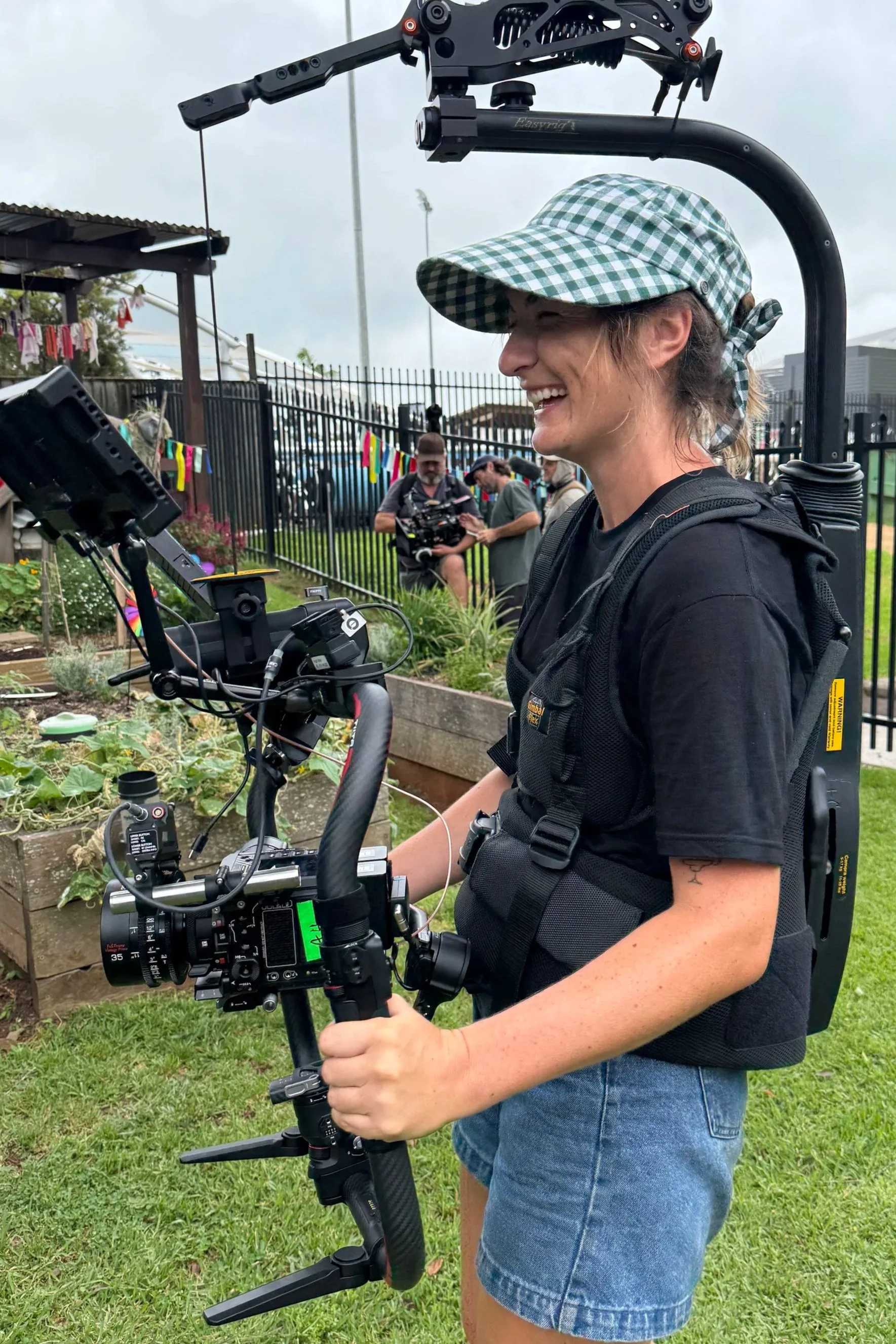 A woman in a green checkered cap and black t-shirt smiling while operating a professional camera mounted on a stabilizer. She is outdoors in a garden with a fence and other people in the background, some with cameras, and colorful decorations hanging.