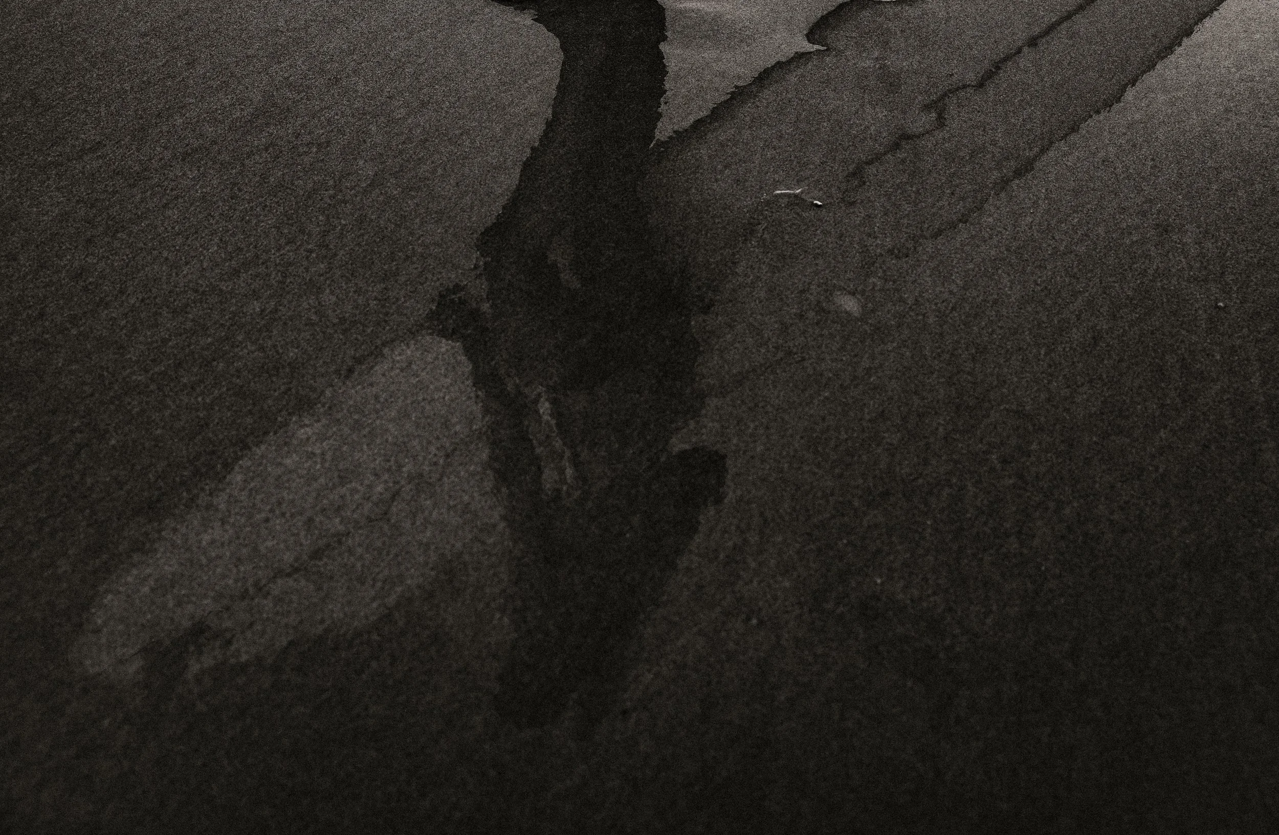 Close-up of dark, wet sand with a small ripple of water, possibly on a beach at night or in low light conditions.