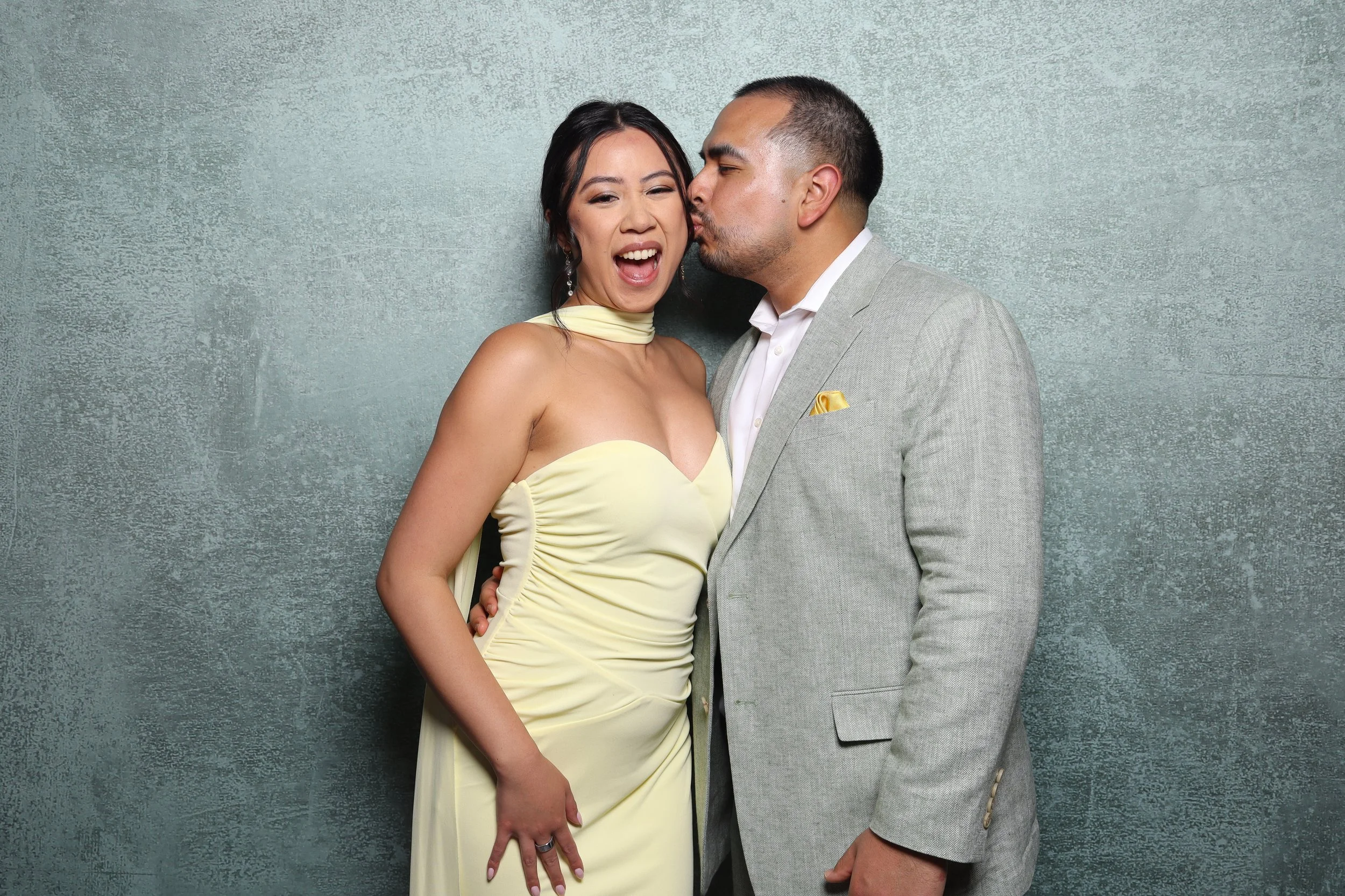 A woman in a yellow dress being kissed on the cheek by a man in a light gray suit against a textured gray background.
