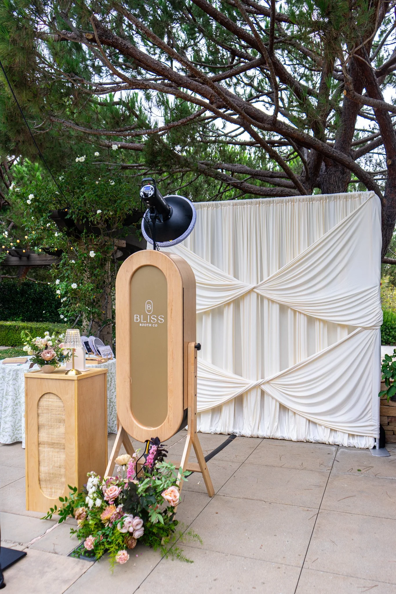Photo of an outdoor photo booth with a draped white curtain backdrop, a light wood frame, and a camera mounted at the top left. There is a small table with a floral arrangement and a lamp to the left of the photo booth. The scene is set on a patio with a large tree overhead and surrounding greenery.