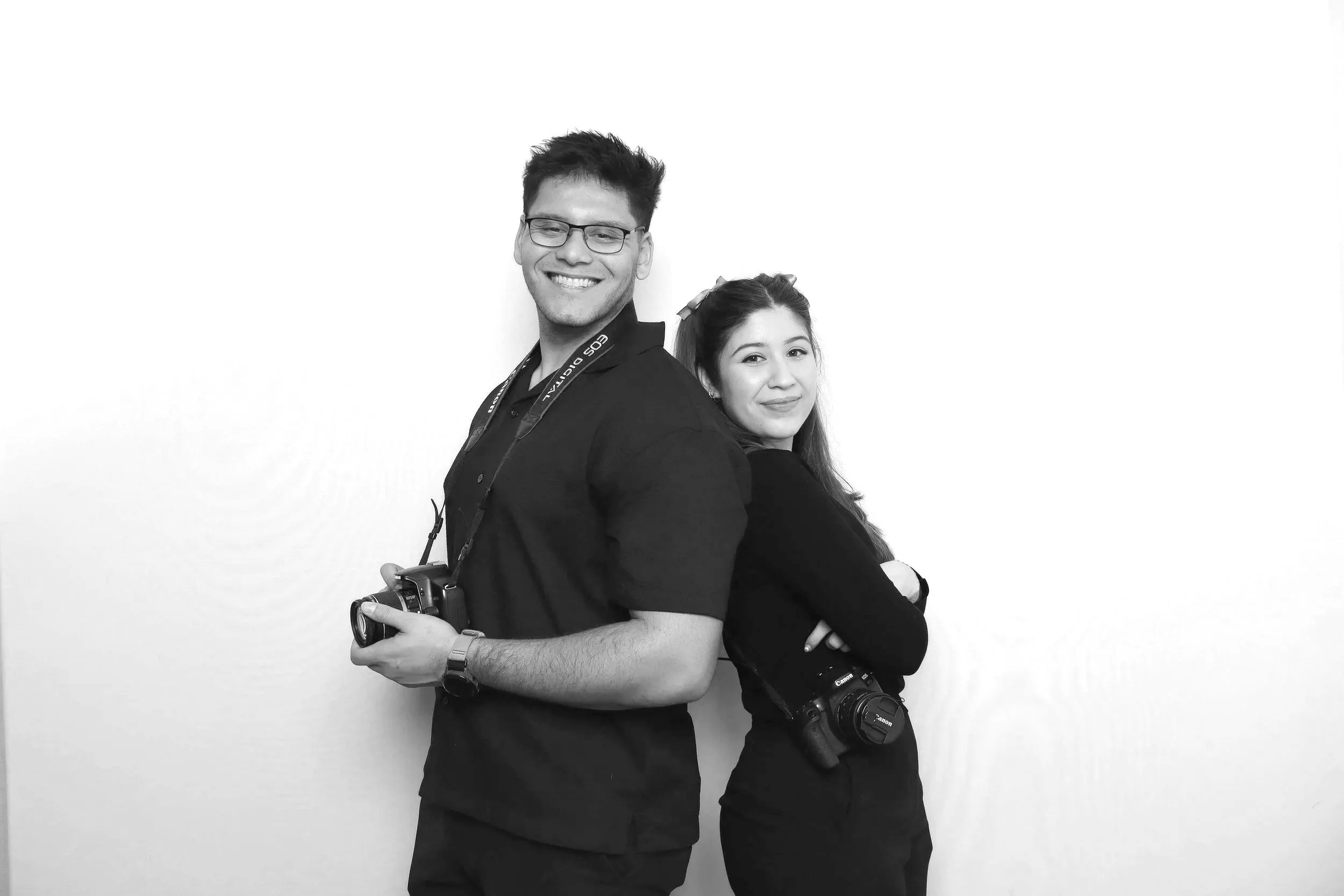 A young man and woman standing back to back, both holding cameras, smiling, against a plain white background.