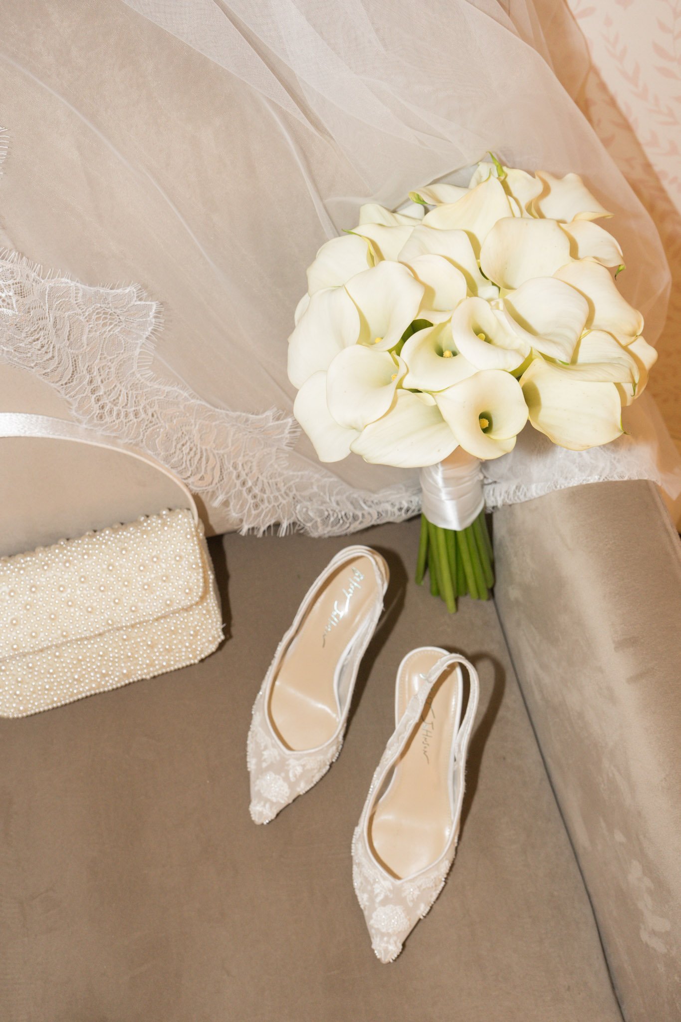 A bouquet of white calla lilies, a pair of white lace high heel shoes, a pearl-embellished clutch, and a cream-colored fabric with lace details on a gray cushioned surface, likely part of a wedding setup.