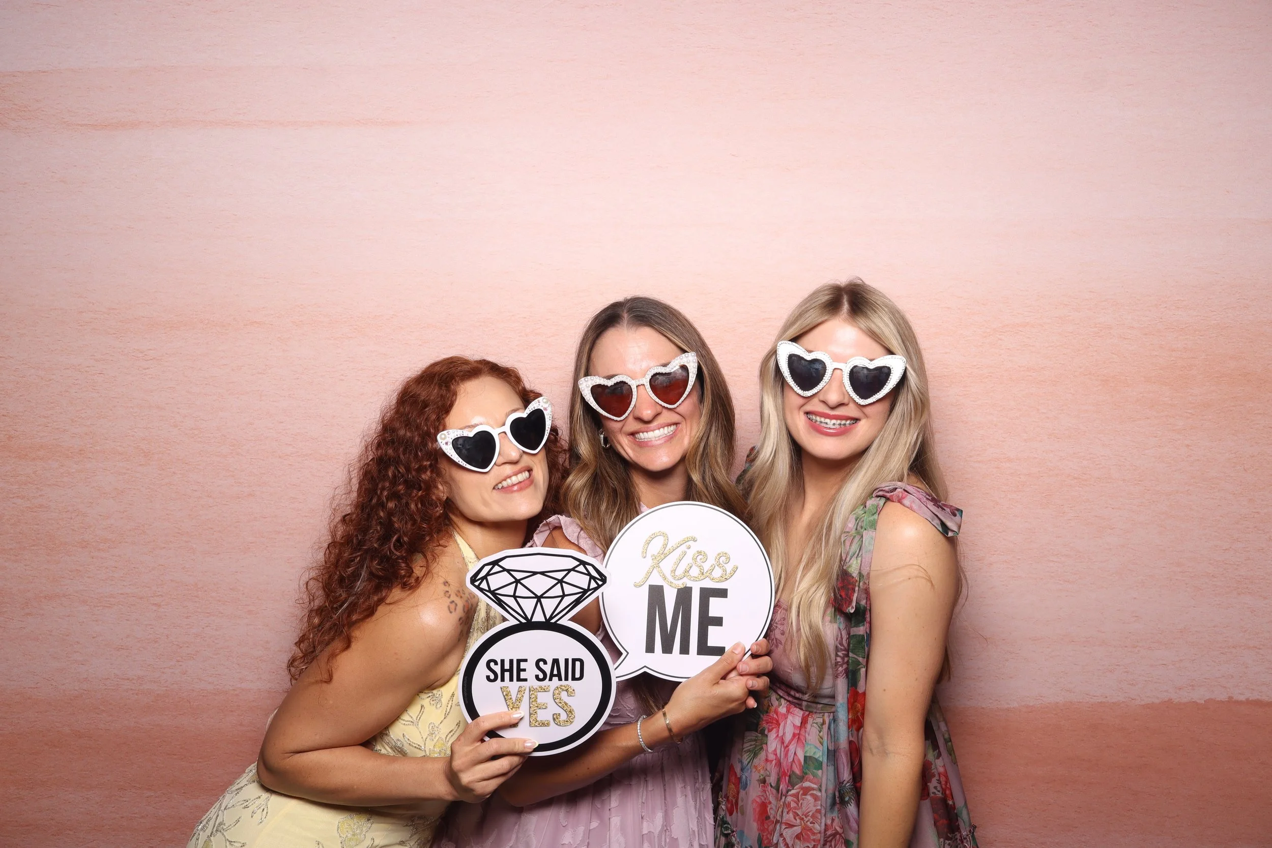 Three women wearing heart-shaped sunglasses smiling, holding signs that say "She Said Yes" and "Kiss Me," against a pink backdrop.