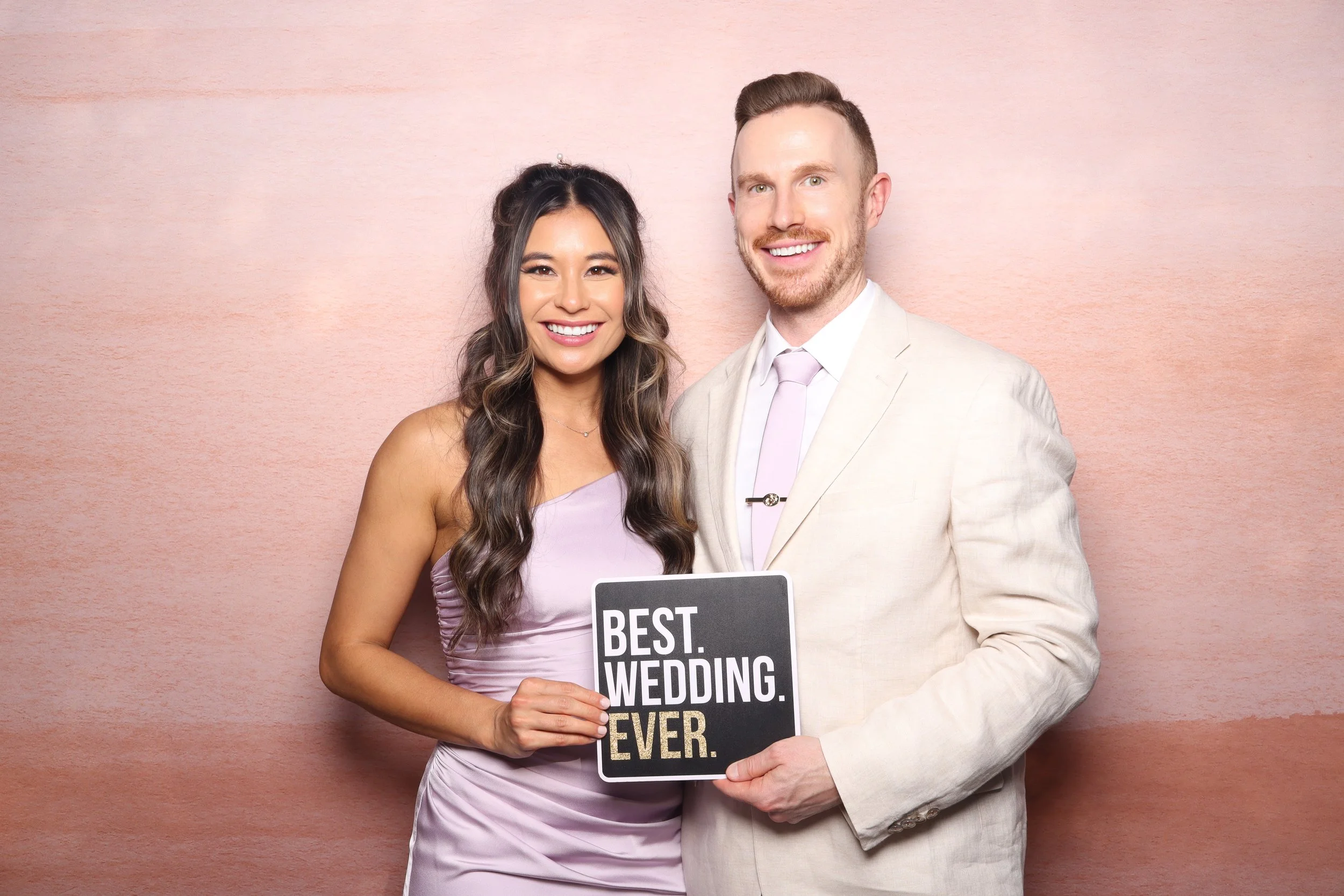 A smiling woman holding a sign that says "Best Wedding Ever" while standing next to a smiling man in a light-colored suit against a pink background.