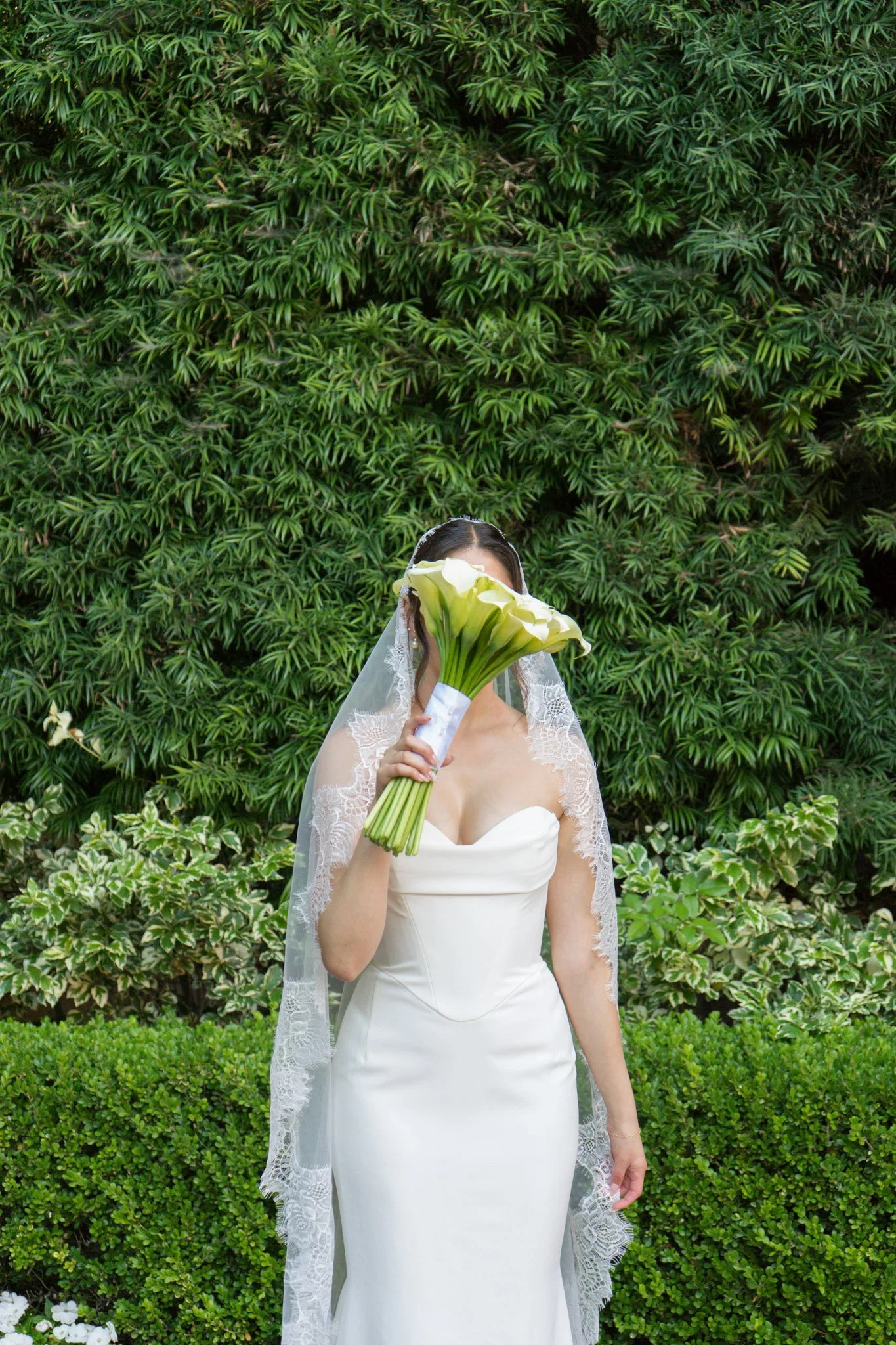 Bride in wedding dress holding a bouquet of calla lilies covering her face, standing outdoors with a green leafy background.