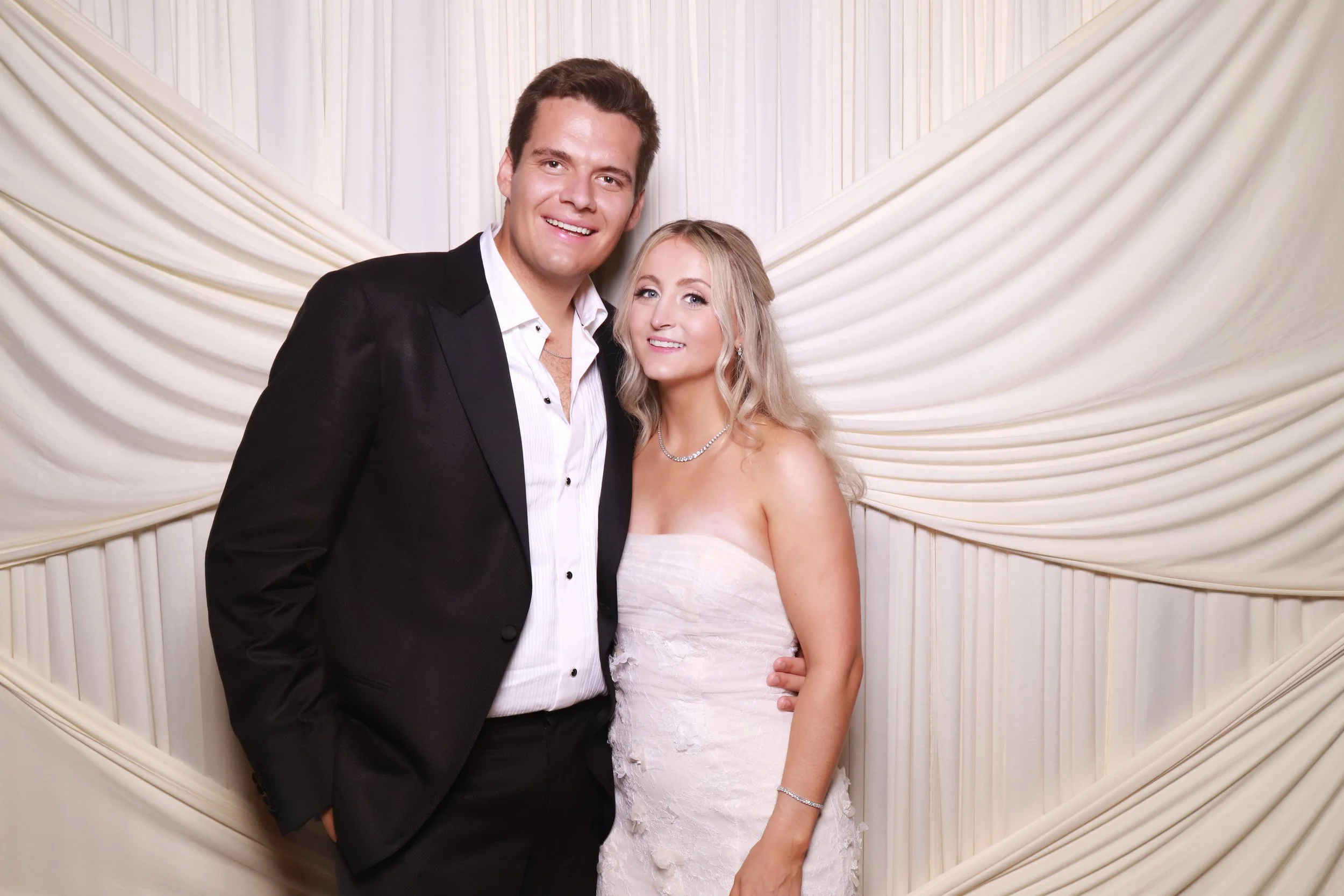 A smiling man and woman dressed in formal attire posing together. The man is in a black suit with a white shirt and the woman is in a strapless white dress with jewelry. They are standing in front of a cream-colored, draped backdrop.