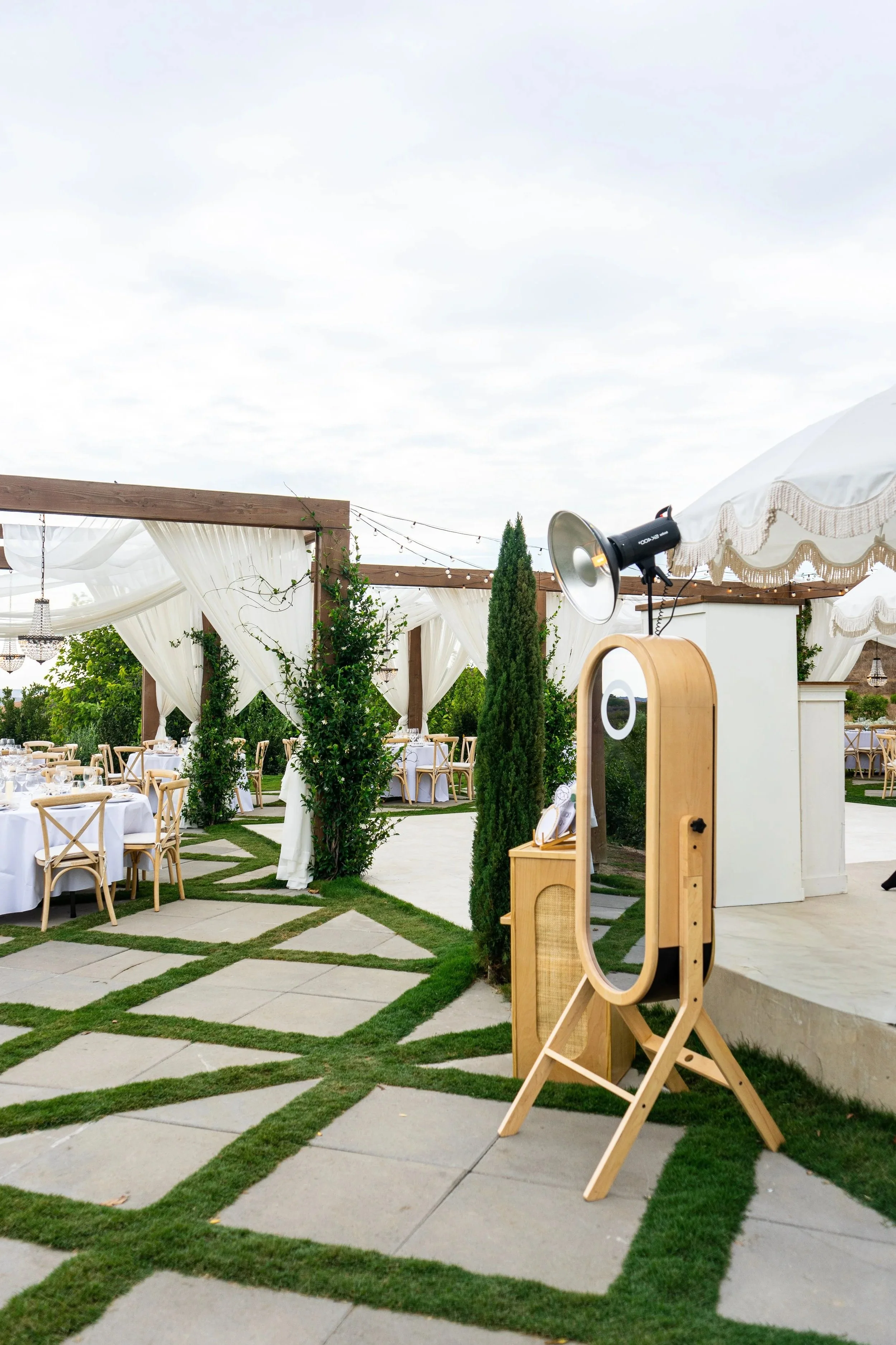 Outdoor wedding or event setup with draped white curtains, wooden chairs, and tables set for dining, decorated with greenery and string lights, and a photography/photo booth area with a large mirror and studio light.