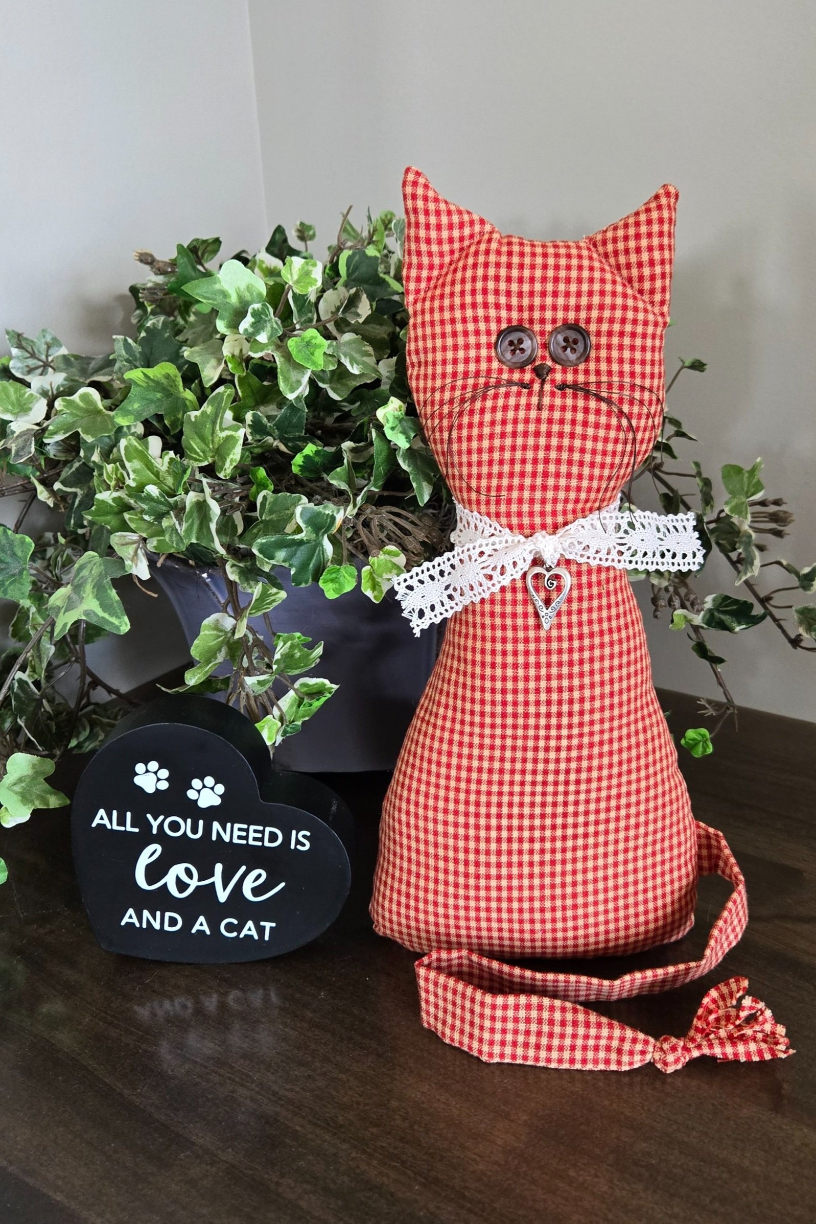 A handmade red and white gingham cat doll with a white crochet lace bow and a silver heart charm. The cat has black button eyes and a black embroidered nose and whiskers.