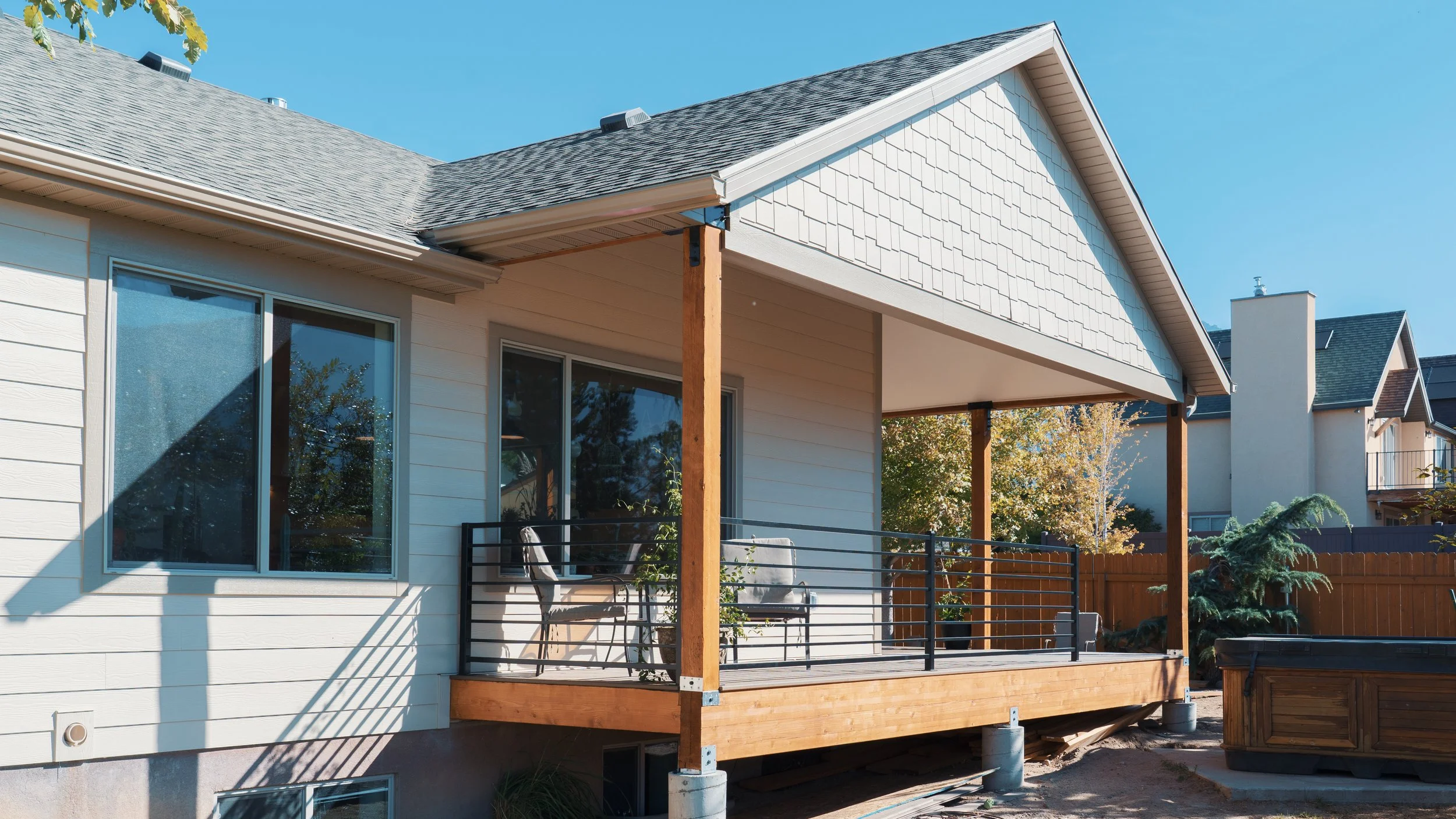 Newly built backyard deck with black metal railings and outdoor furniture, attached to a white house with large windows, in a suburban neighborhood under a clear blue sky.