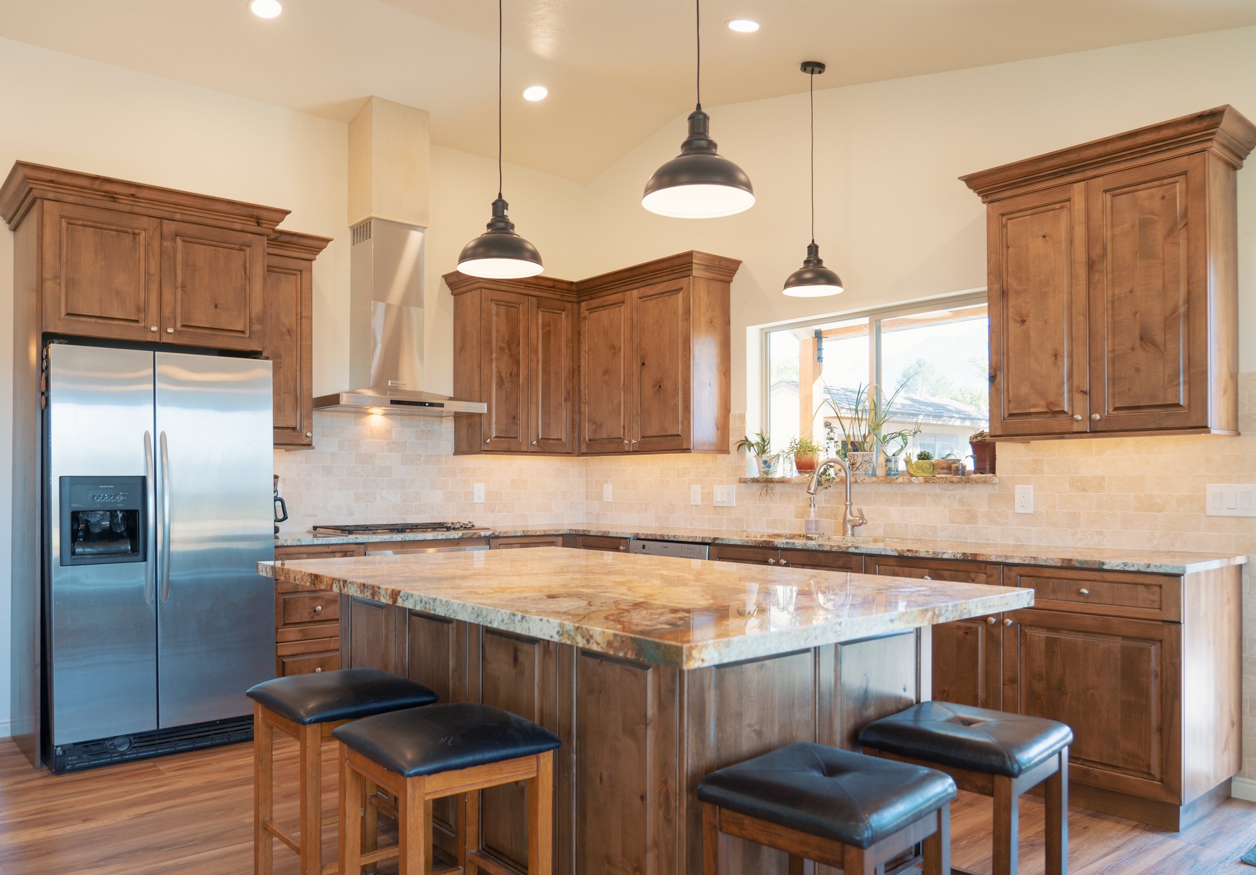 Modern kitchen with wooden cabinets, granite island, stainless steel refrigerator, and pendant lighting.