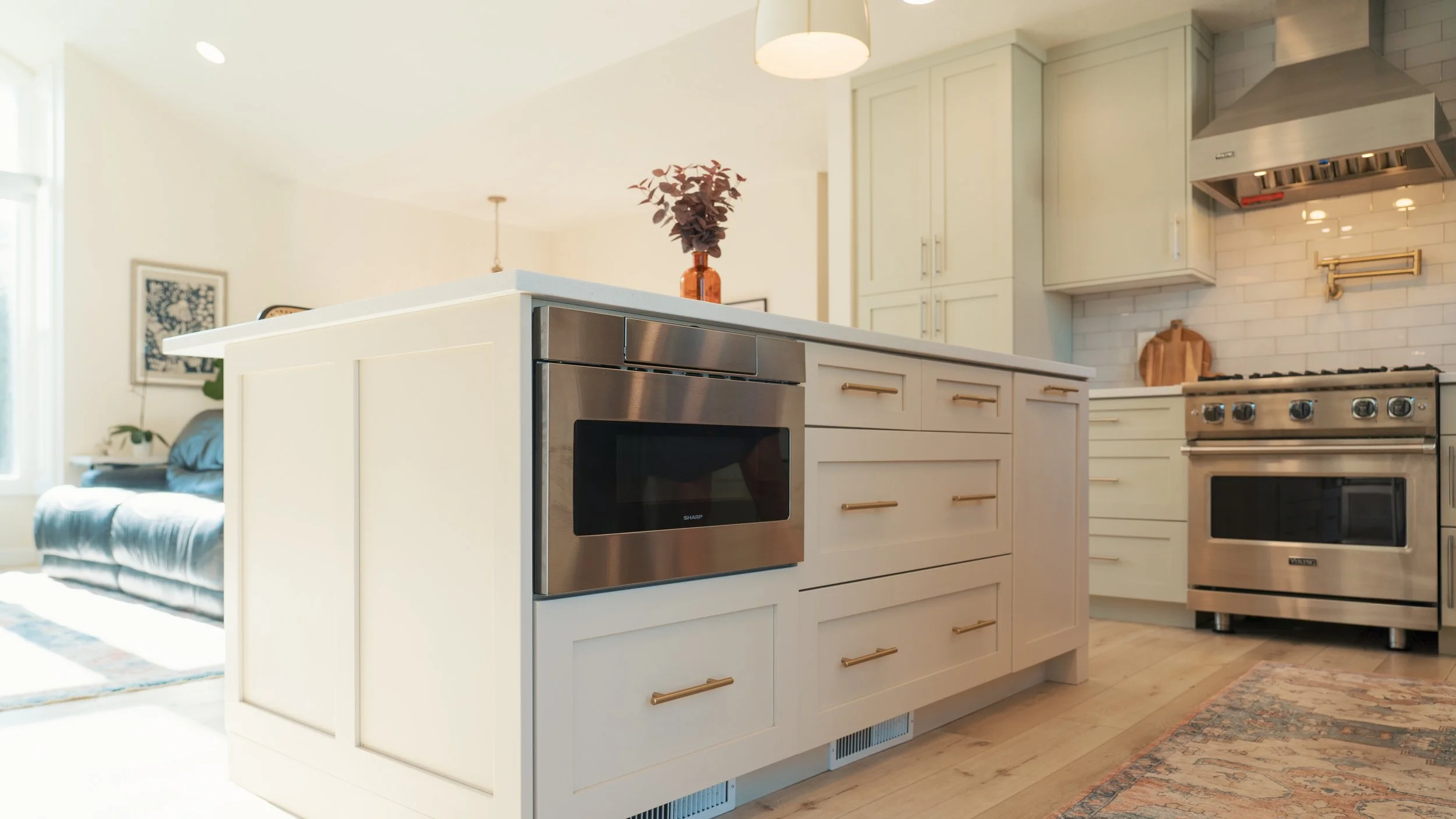 Modern kitchen with white cabinetry, stainless steel appliances, a kitchen island with a microwave, and a living room area with a sofa visible in the background.