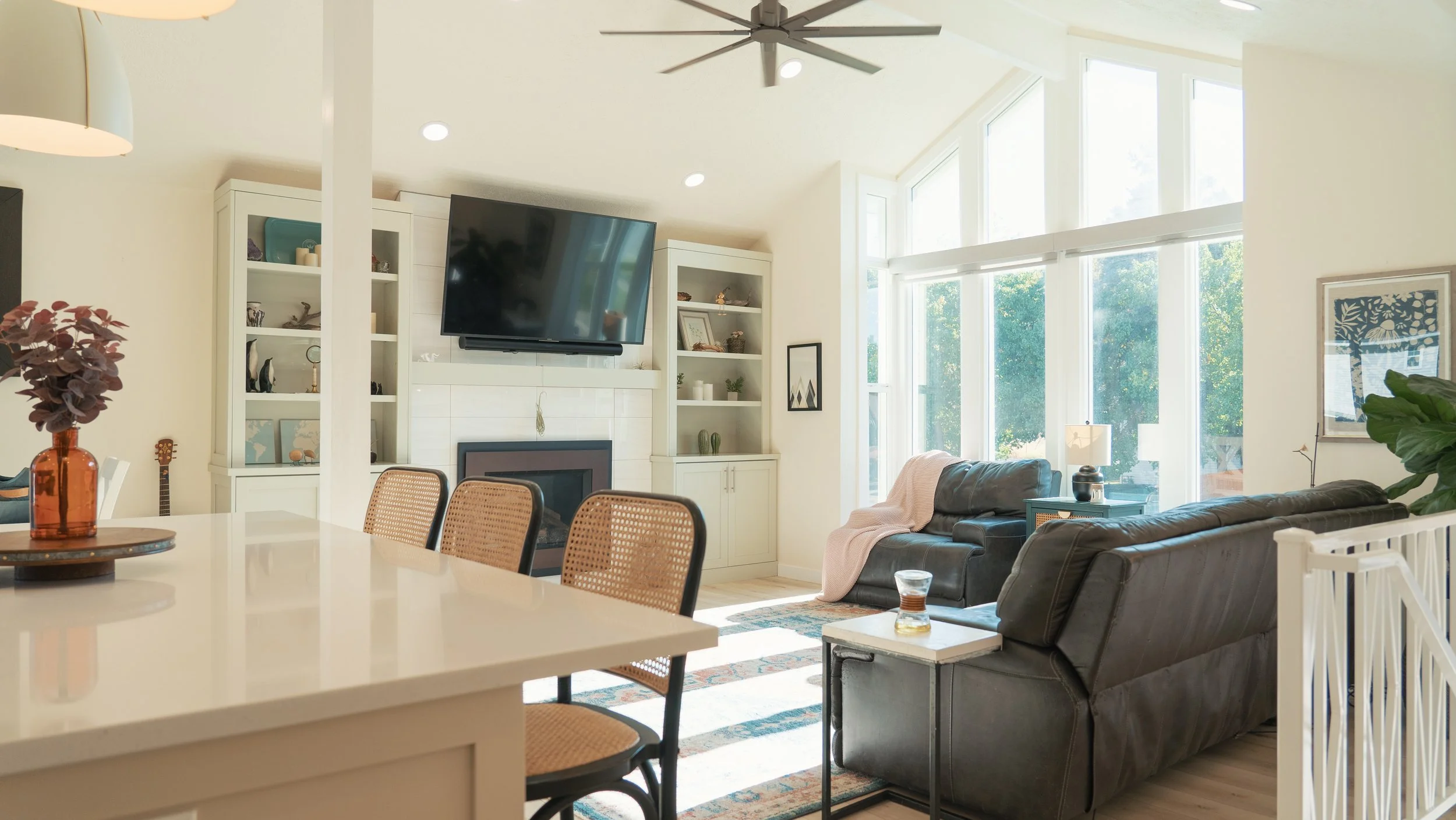 Bright living room with large windows, two black leather couches, a flat-screen TV mounted above a white fireplace, light-colored built-in shelves, and a colorful area rug.