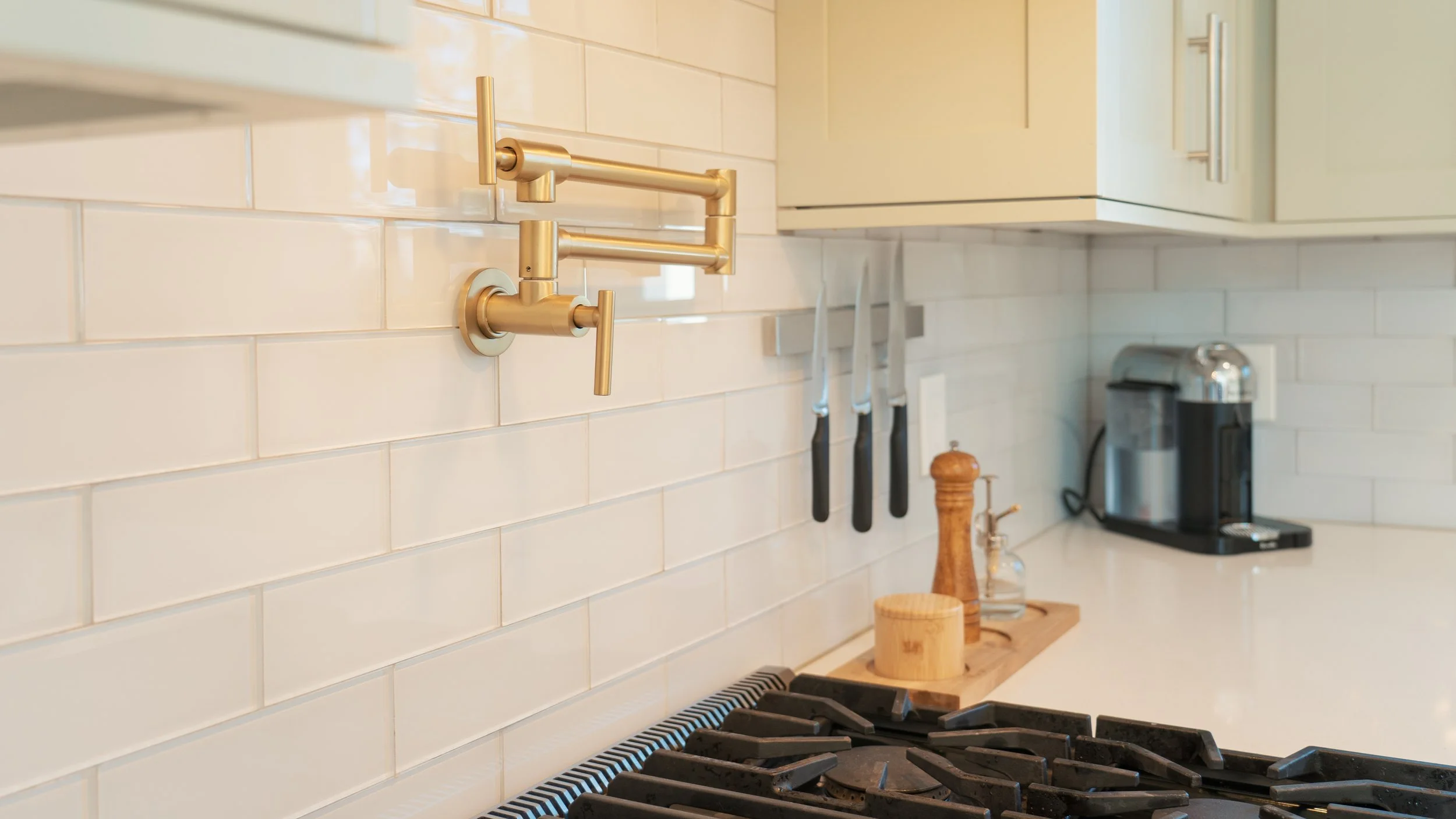 A modern kitchen with a brass faucet mounted on a white subway tile backsplash, a gas stove with black grates, knives hanging on a magnetic strip, a pepper grinder, a wooden cutting board, and a coffee maker on the white countertop.