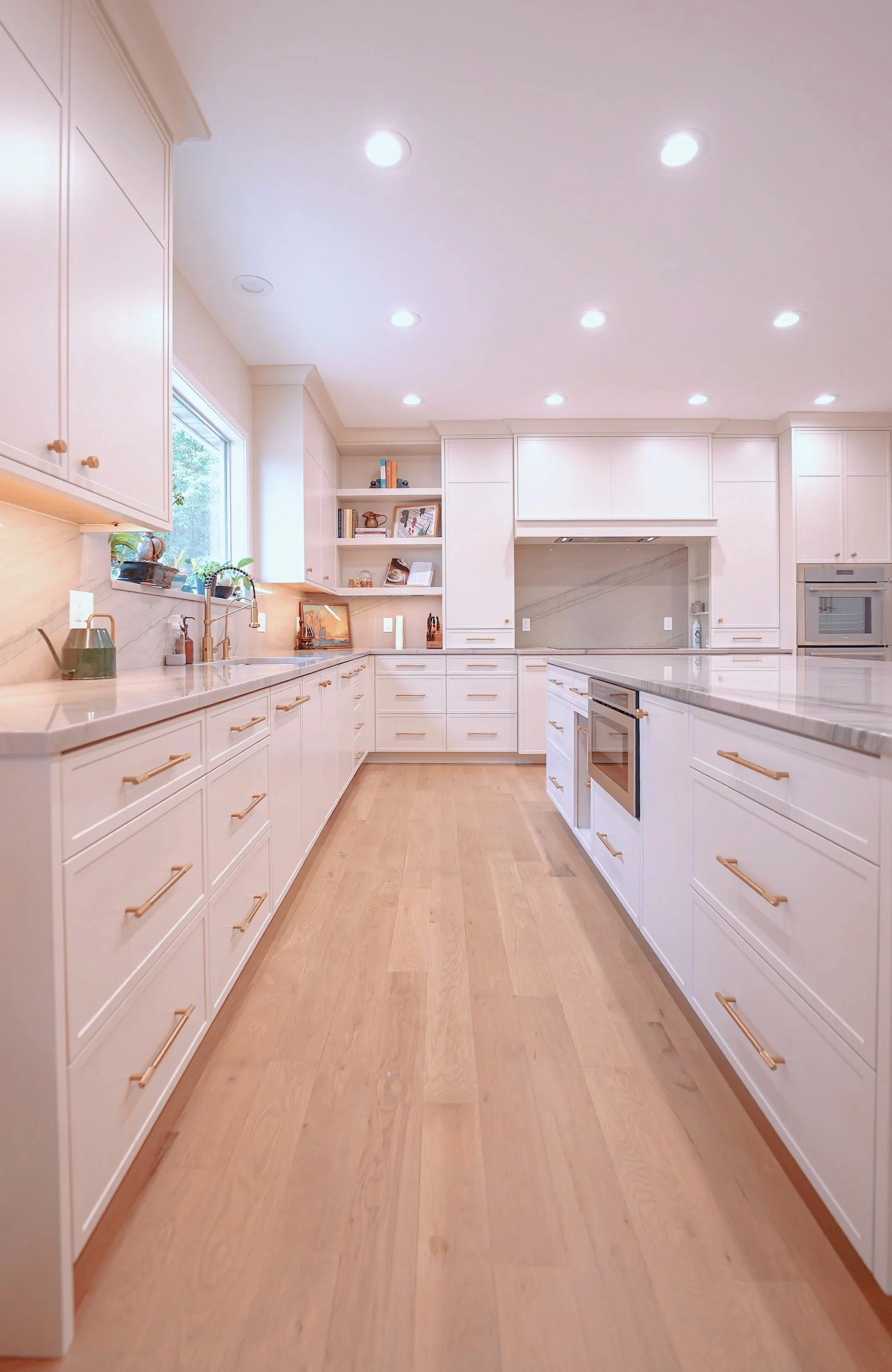 Modern white kitchen with a large island, stainless steel appliances, hanging pendant lights, and a window showing green trees outside.