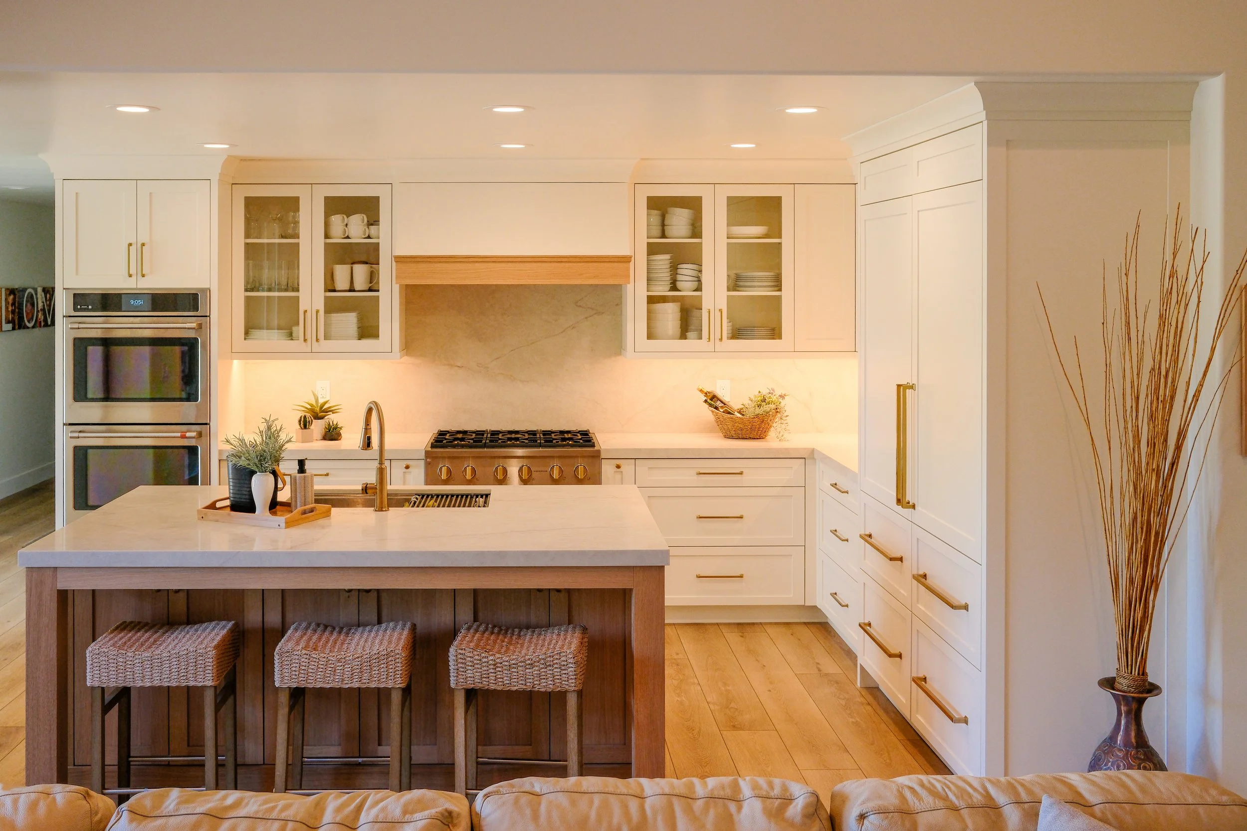 Modern white kitchen with a large island, stainless steel appliances, hanging pendant lights, and a window showing green trees outside.