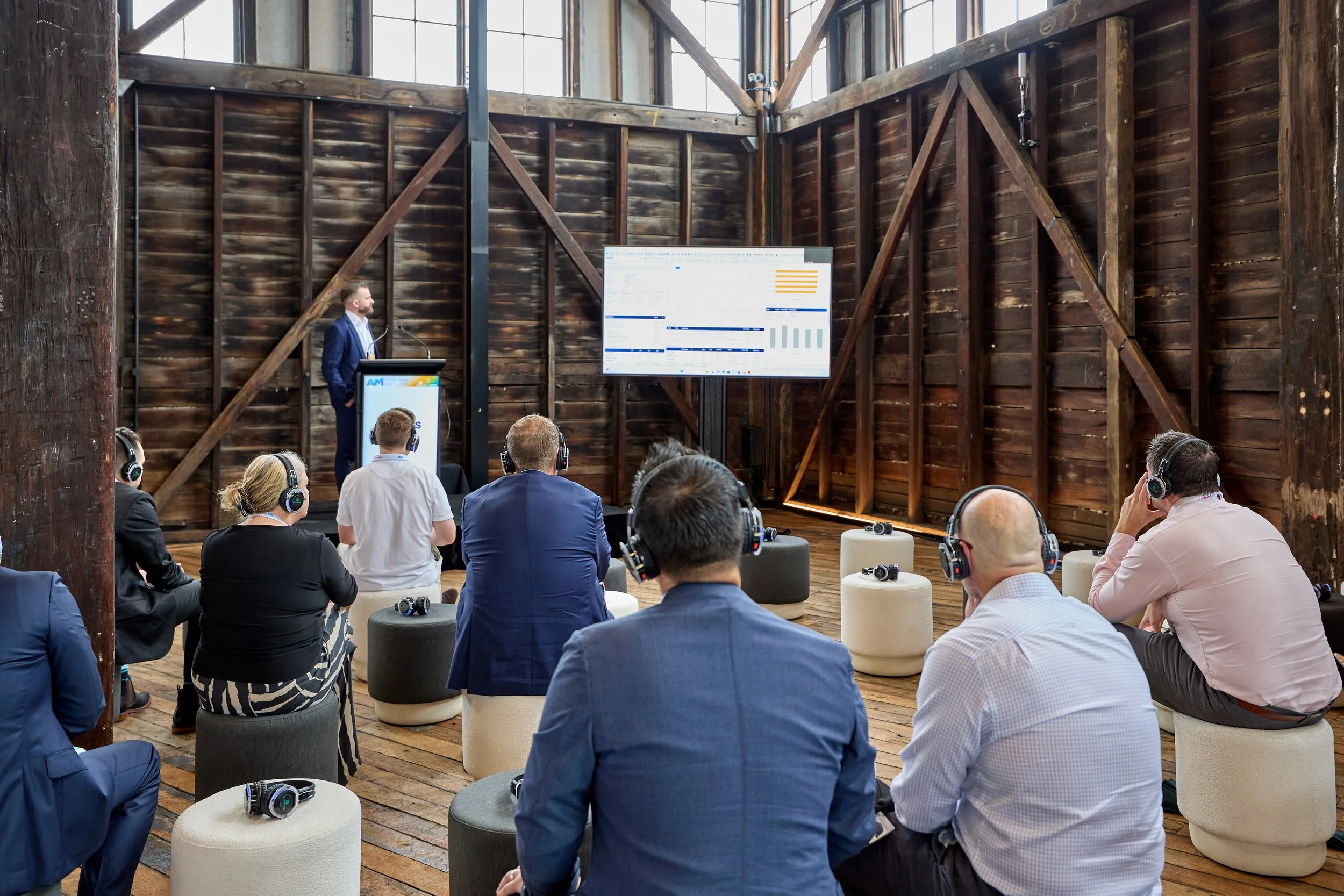 Business presentation in a rustic wooden event space with a speaker at a podium and an audience wearing headsets, watching a large screen displaying charts and graphs.