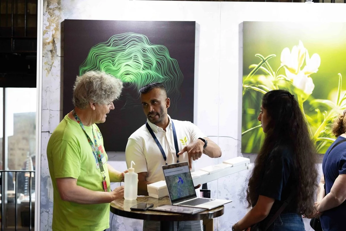 Three people engaged in conversation at an exhibition booth, with a laptop, bottles, and backdrop artwork displaying green topographical and floral images.