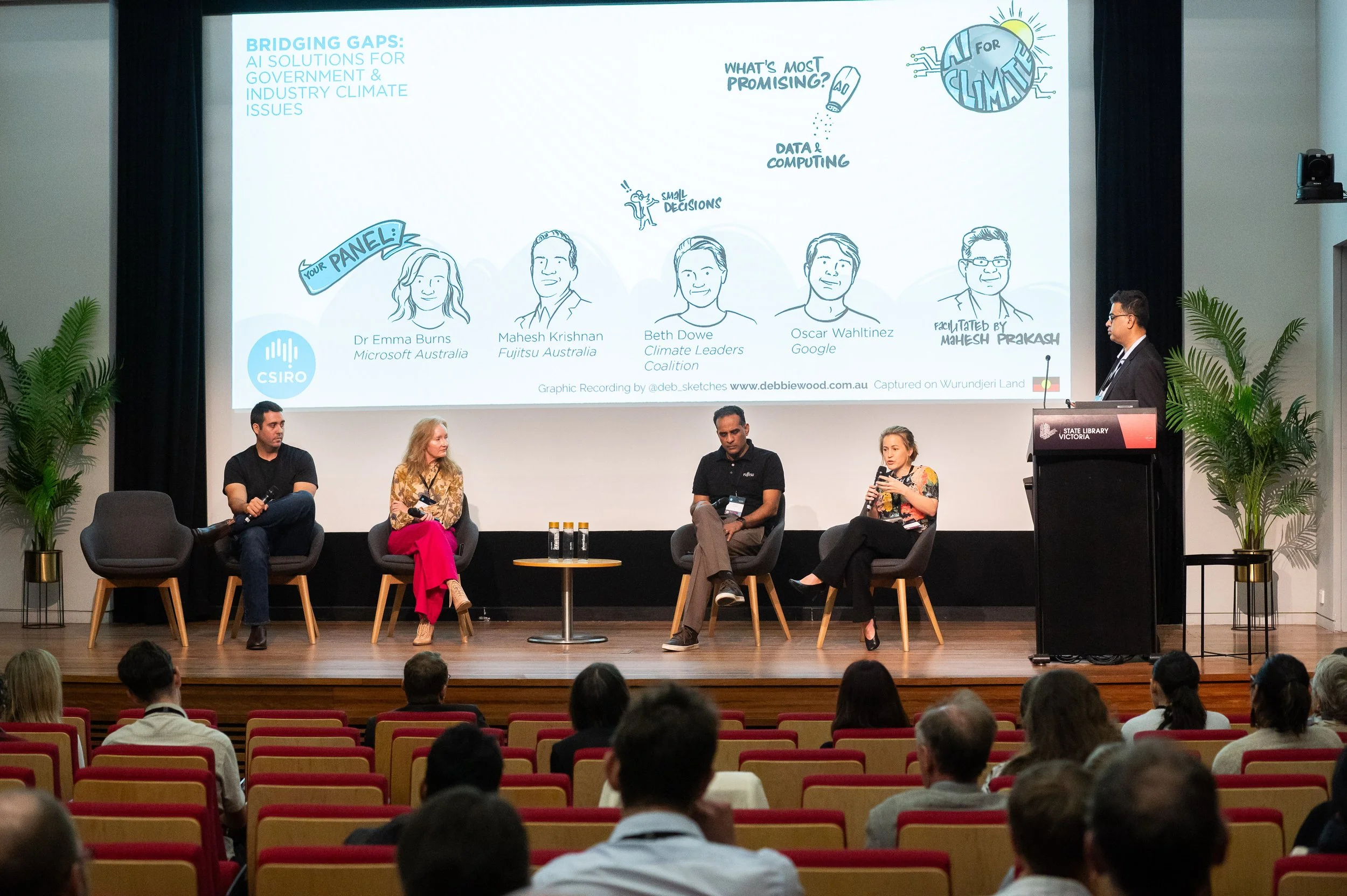Panel discussion at a conference with five individuals seated on stage, one person standing at a podium, large screen behind them displaying a graphic about climate solutions, lush green plants on either side of the stage, and an audience seated in red and beige chairs.