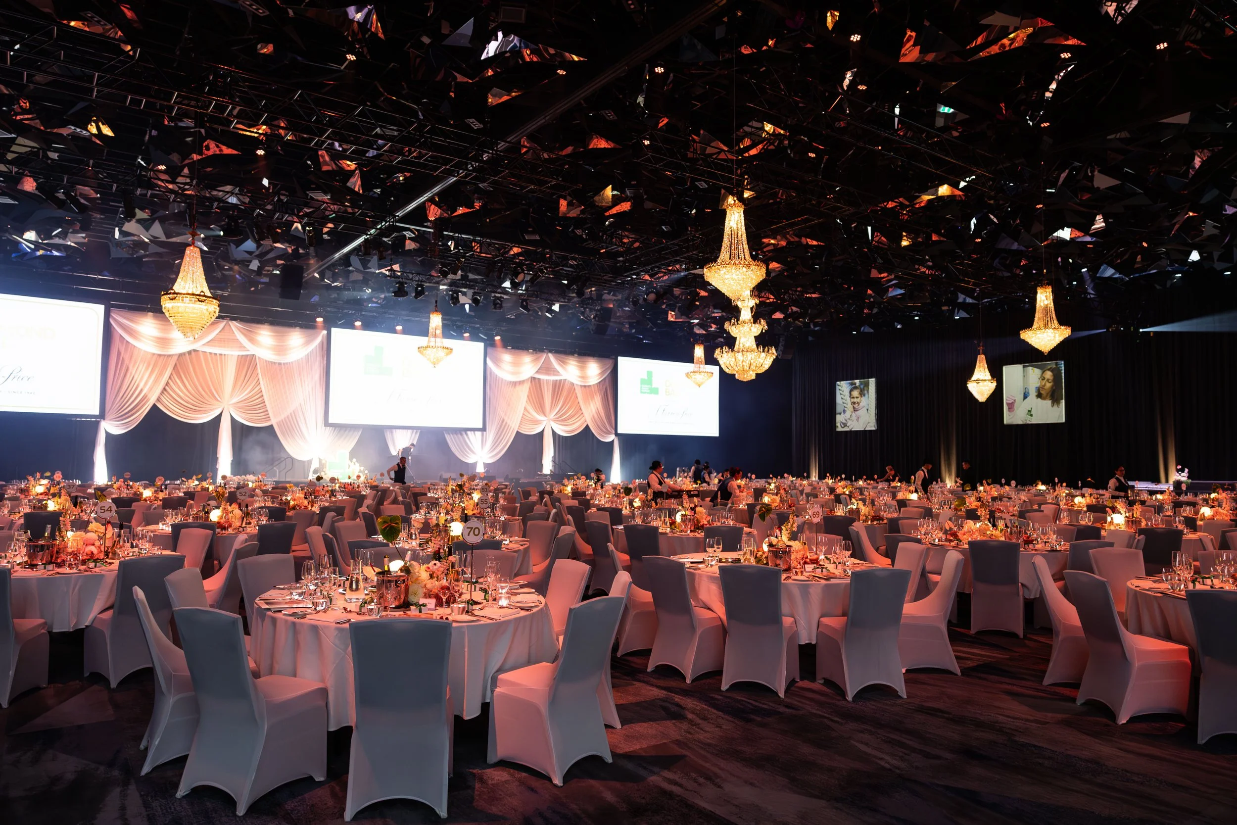 Elegant banquet hall decorated with drapes, chandeliers, and round tables set for a formal event, with large screens and framed photographs on the walls.