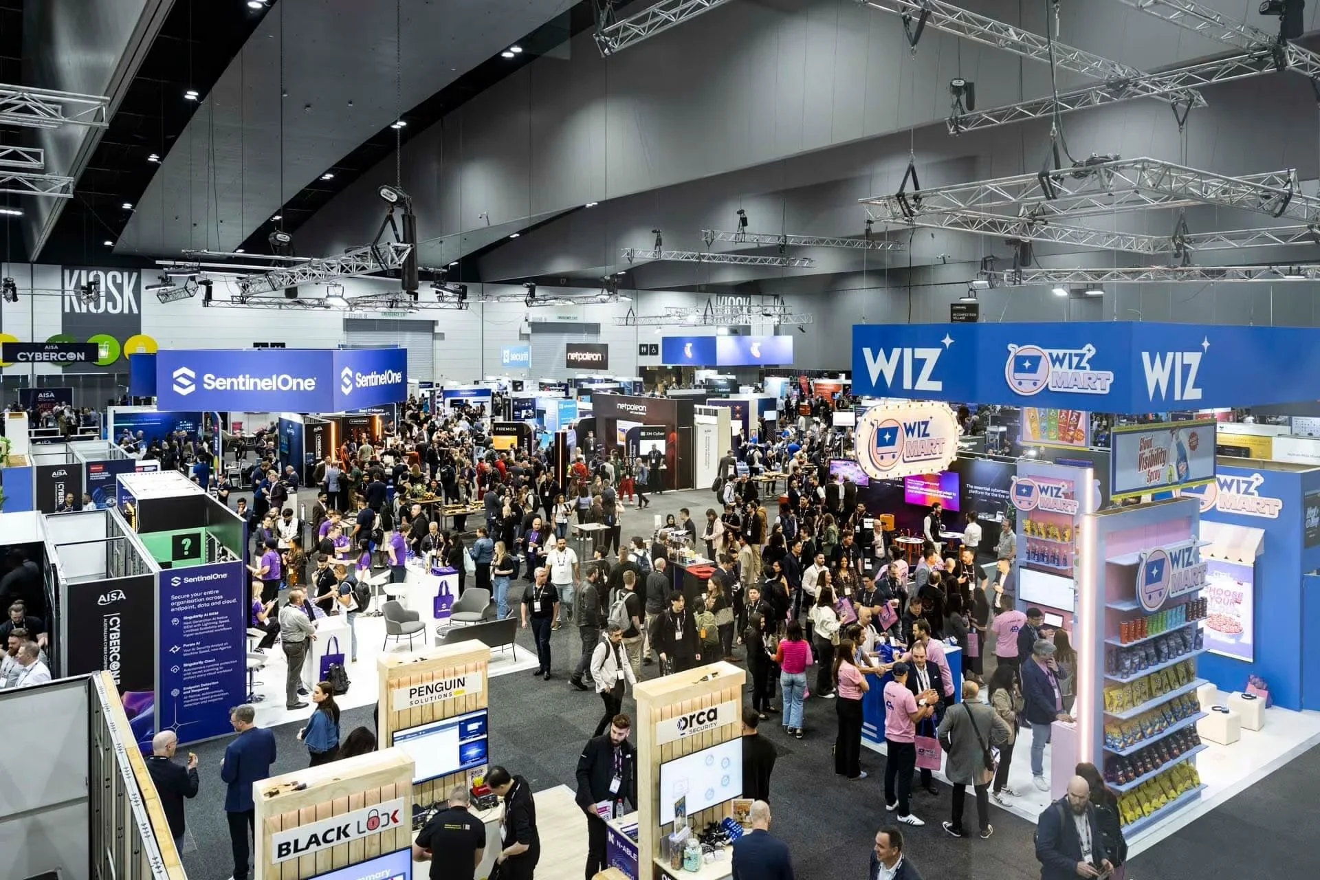 A crowded technology exhibition with various booths and displays from companies like SentinelOne, WIZ, and others, with attendees walking around, exploring exhibits, and engaging with booths.