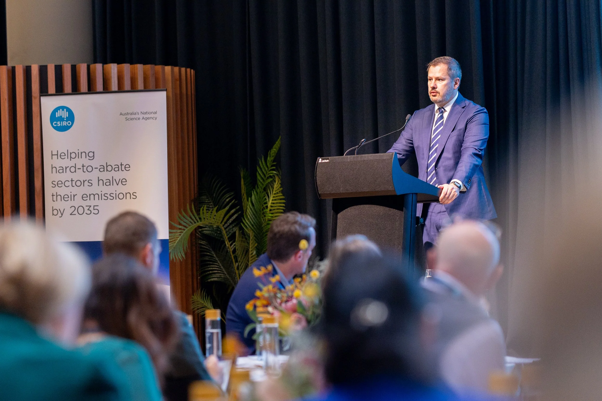 A man in a suit and striped tie is standing at a podium giving a presentation to an audience in a conference room with black curtains behind him. A CSIRO banner nearby Several audience members are seated at tables, with some flowers and water bottles