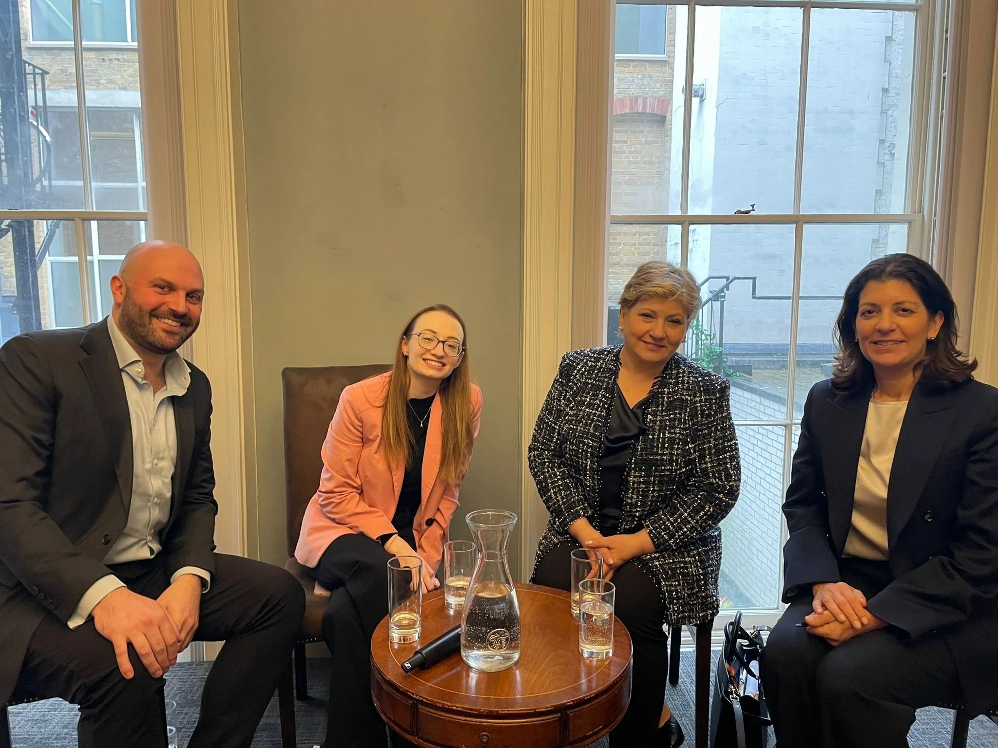 Four people, three women and one man, sitting around a small wooden table in a room with large windows. They are dressed in business attire and smiling at the camera. On the table, there is a water pitcher, glasses, and a microphone.