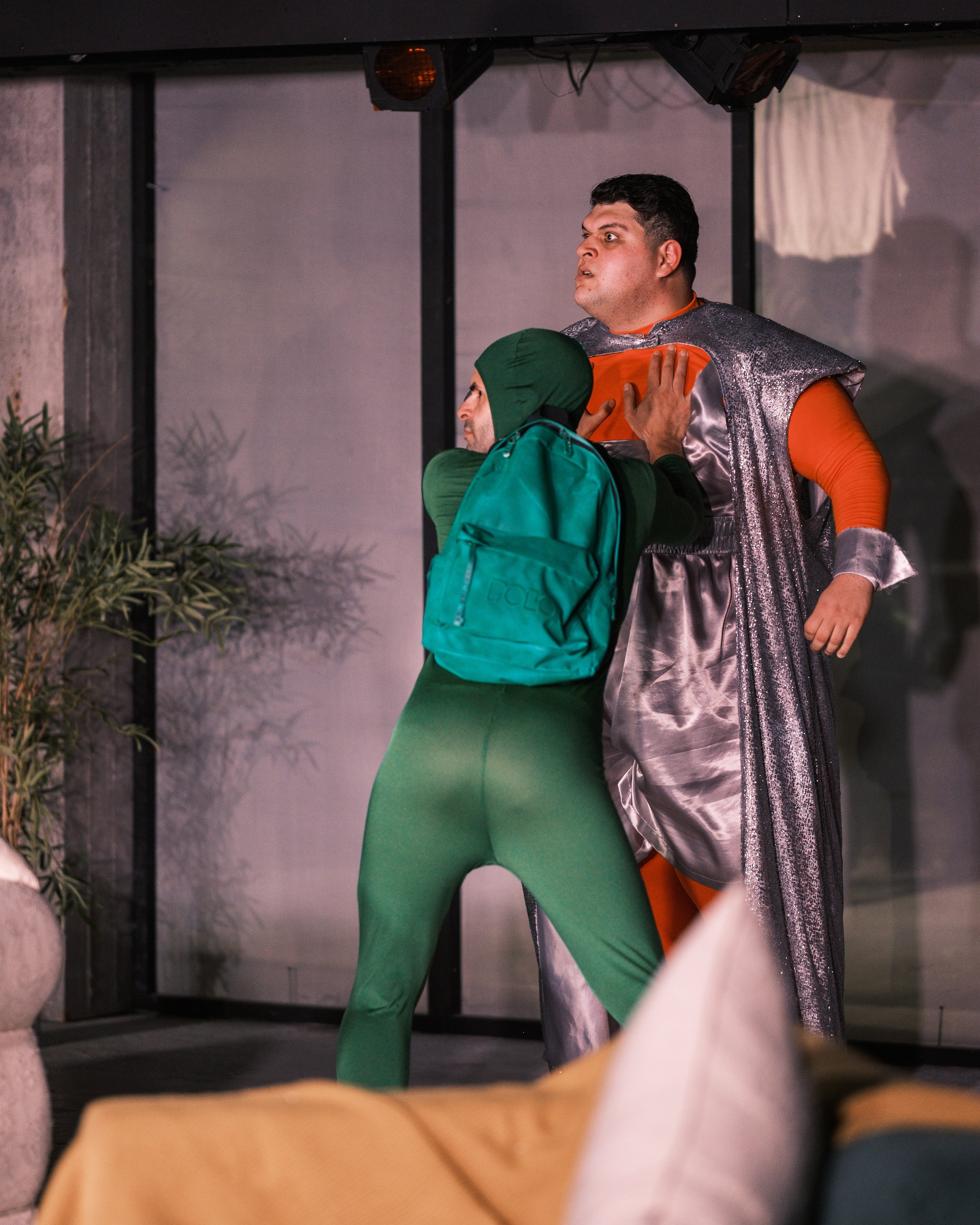 A woman in green attire and a man in a metallic costume appear to fight in a staged scene, with the woman grabbing the man by his shirt. The setting looks like an indoor stage or performance area.