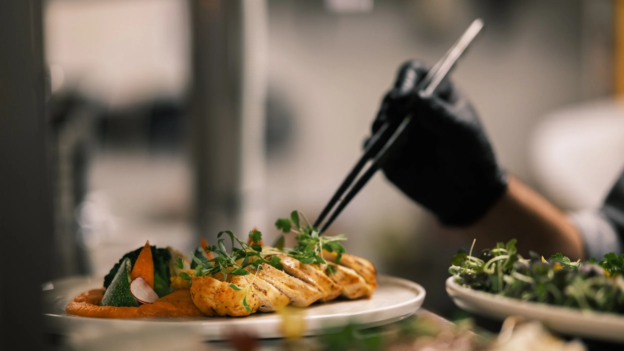 Chef using tongs to place grilled chicken slices with fresh greens and vegetables on a white plate in a professional kitchen.