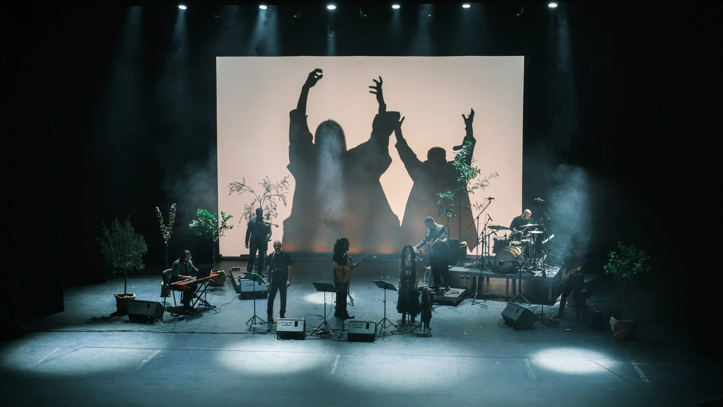 A band performs on stage with a large screen behind them showing silhouettes of three people with their hands raised. The stage has plants and musical instruments, including drums, guitars, a keyboard, and microphones.